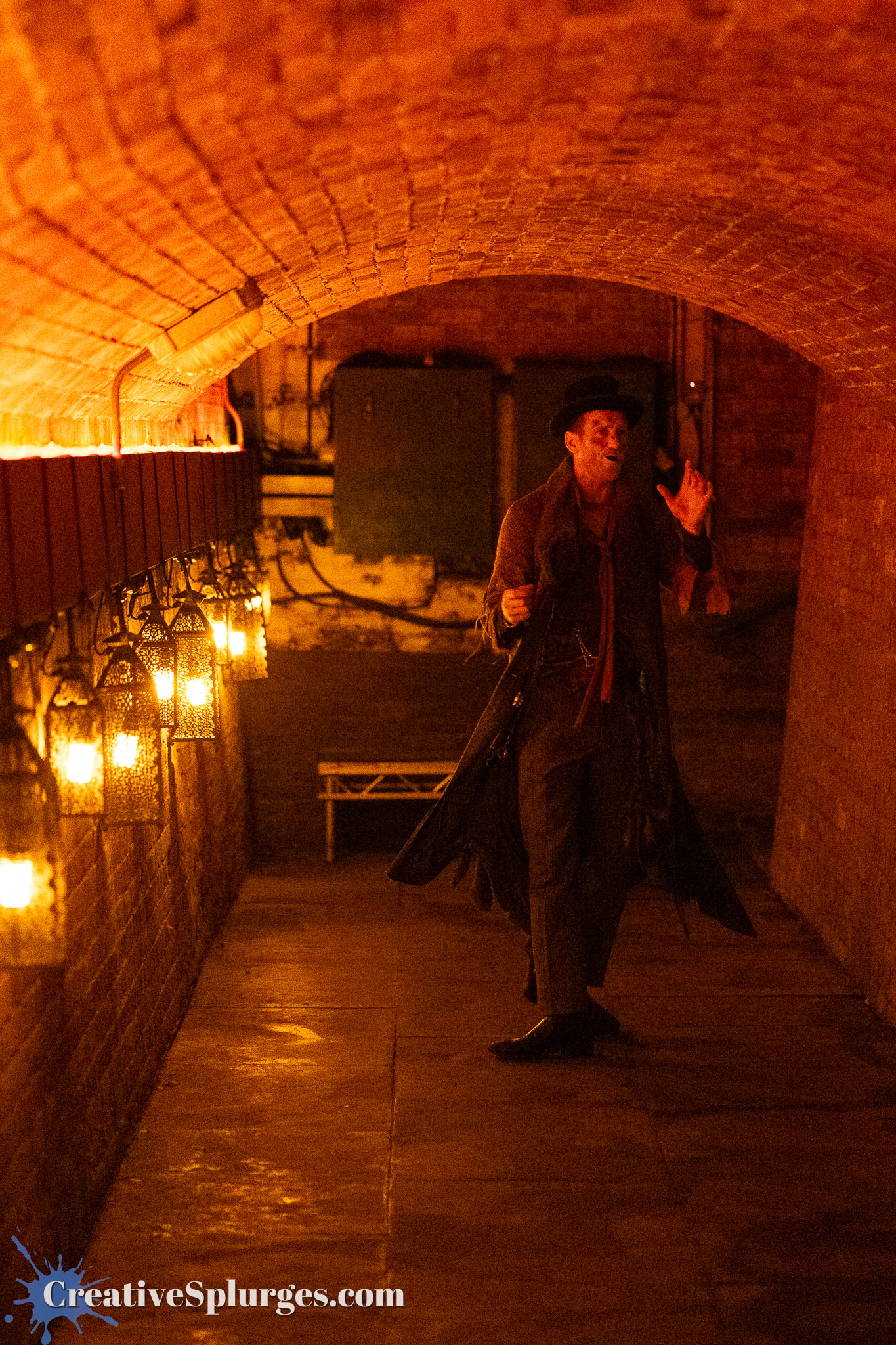 A man in a long coat and hat standing in a dimly lit brick tunnel lined with lanterns, gesturing as if speaking or performing.
