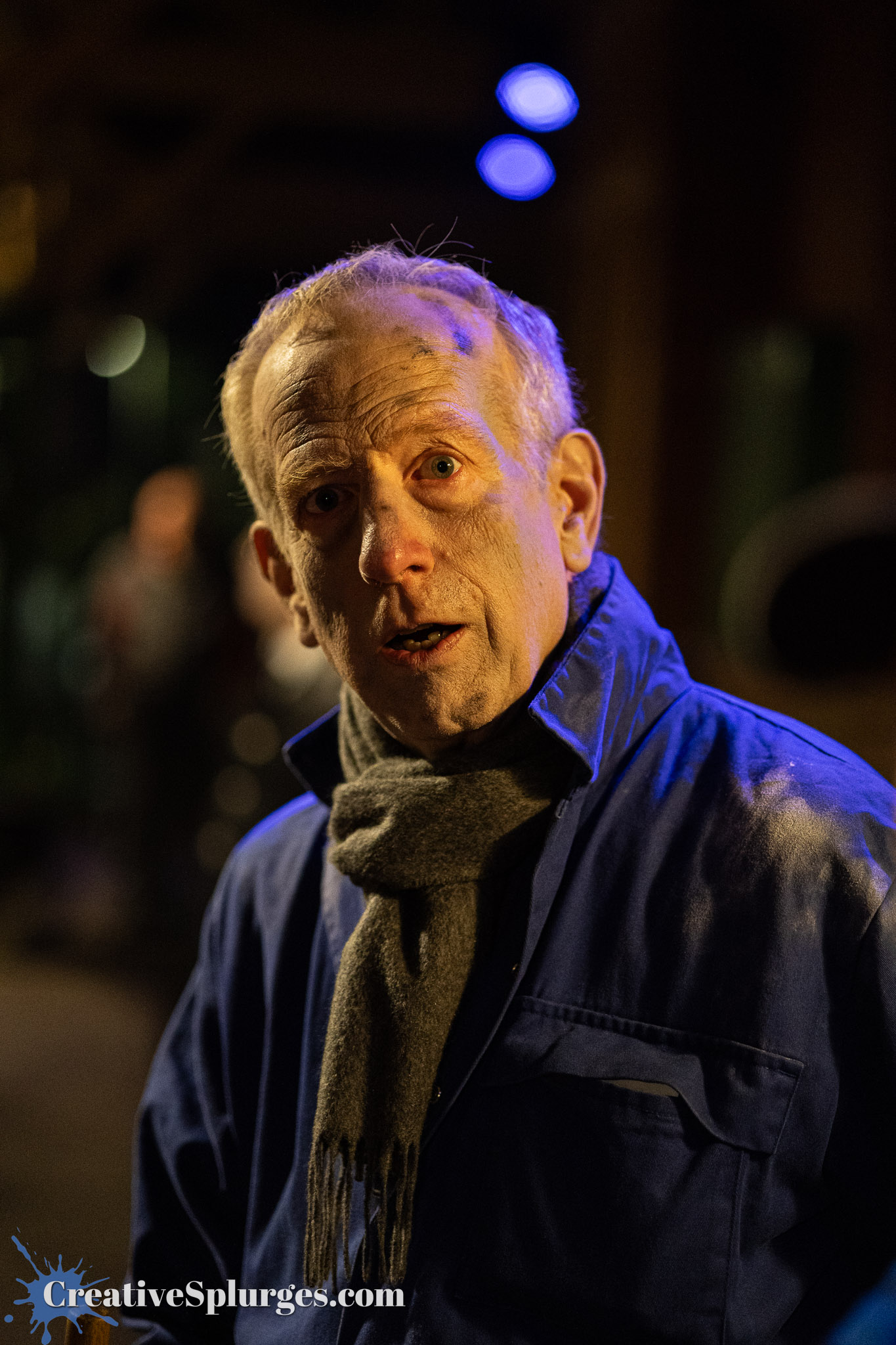 A man with graying hair and a scarf, looking surprised or concerned, illuminated by soft blue lighting in a dimly lit environment.