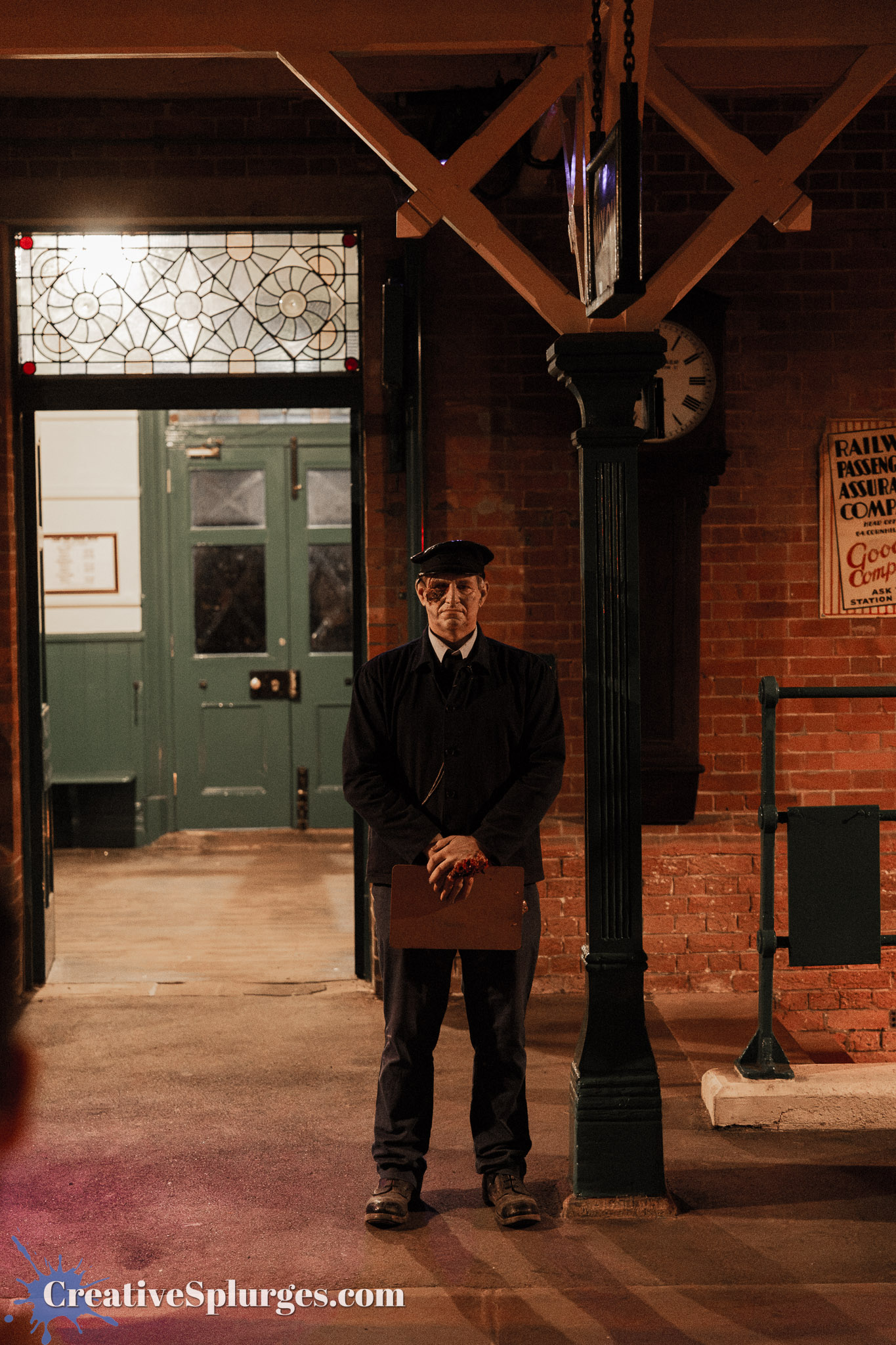 A station attendant standing in a vintage train station, holding a clipboard, wearing a dark uniform and a cap, with red brick walls and green doors in the background.