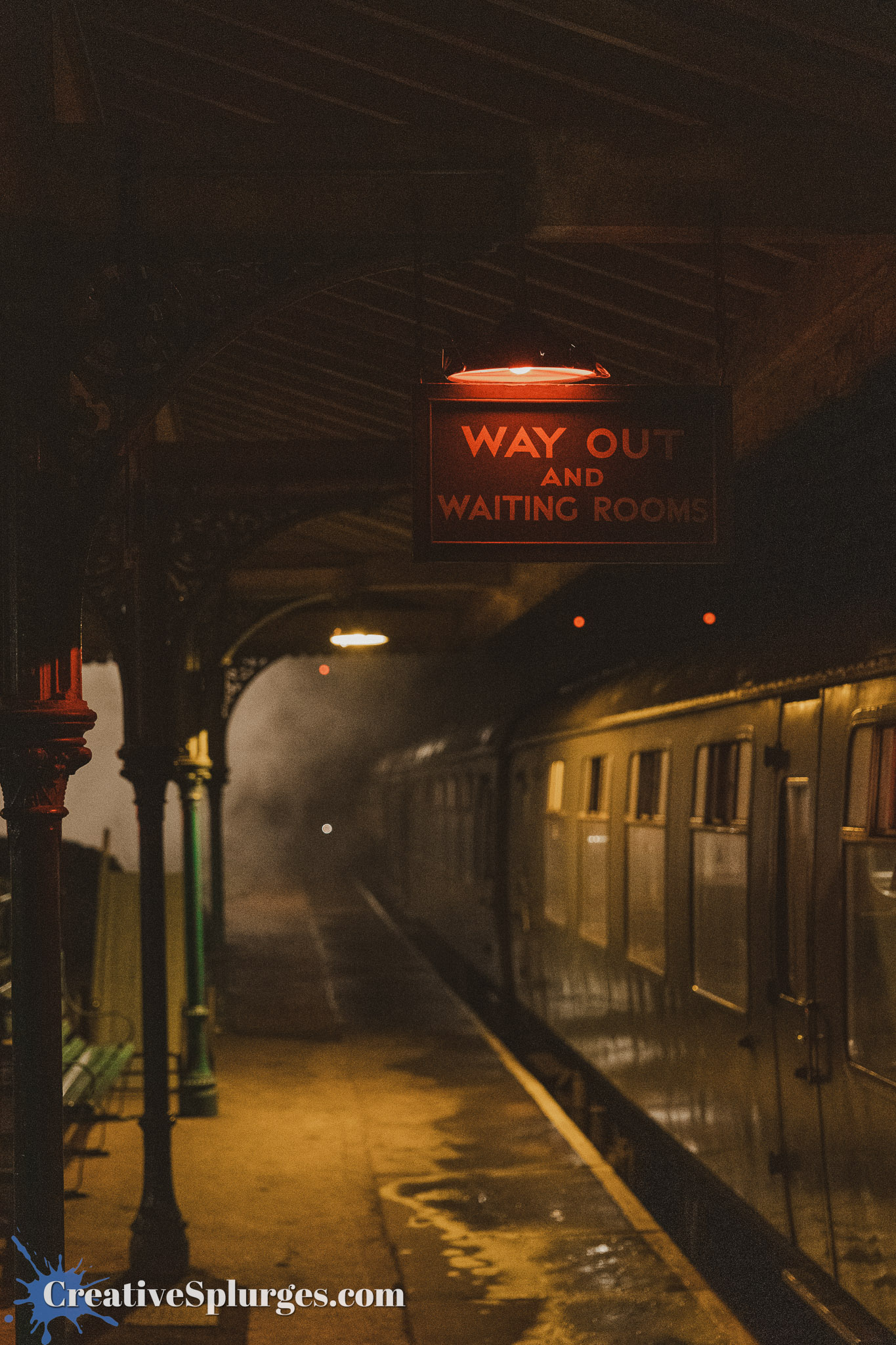 A dimly lit train station platform featuring a sign that reads 'WAY OUT AND WAITING ROOMS,' with steam and a vintage train visible in the background.