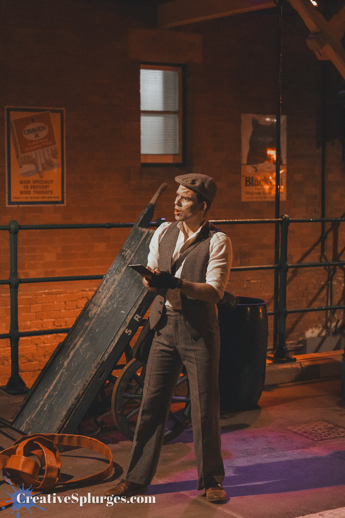 A performer in vintage attire, including a flat cap and vest, holds a clipboard while standing in a dimly lit room with exposed brick walls and various props, including an old cart and signage.