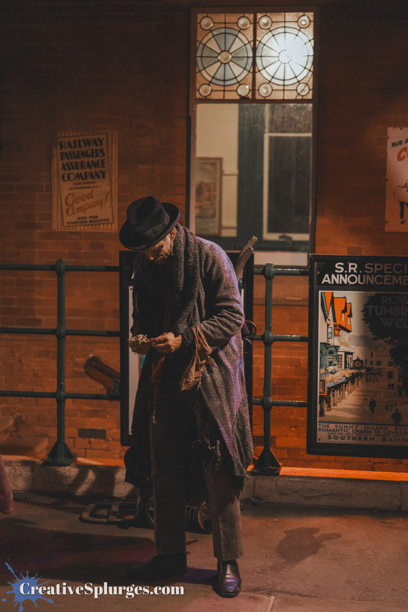 A man dressed in period clothing stands under a dimly lit building, holding a small object in his hands. He wears a hat and a long coat, with a backdrop featuring old advertising posters and stained glass windows.