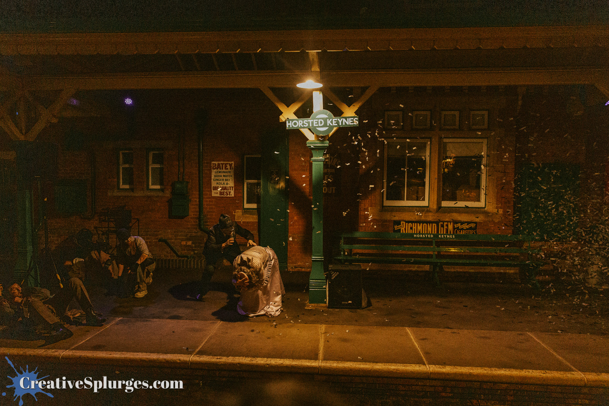 A nighttime scene at Horsted Keynes train station, featuring a historical platform with a sign, a bench, and several people sitting and interacting, while paper confetti floats through the air.