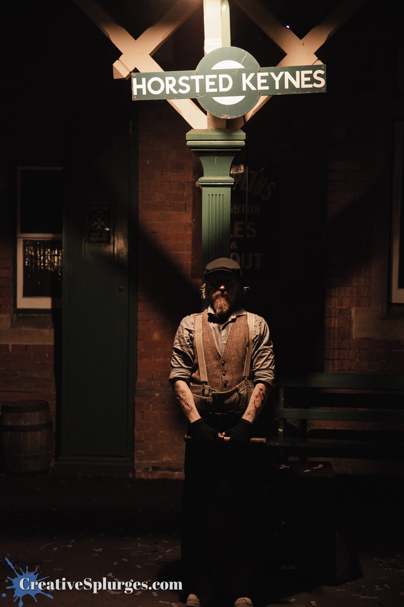 An individual dressed in vintage clothing stands at a dimly lit train station sign that reads 'Horsted Keynes.' The scene is set at night with a rustic background and the person holding a cane, illuminated by soft lighting.