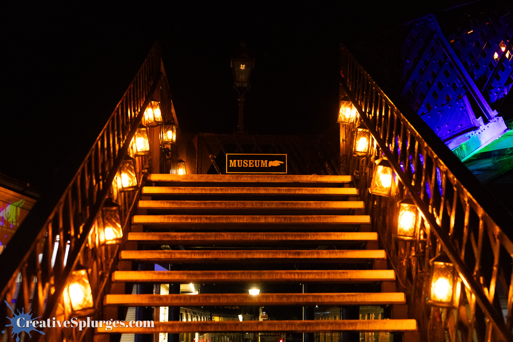A staircase illuminated by warm lights leading to a sign that reads 'MUSEUM', set against a dark night sky.