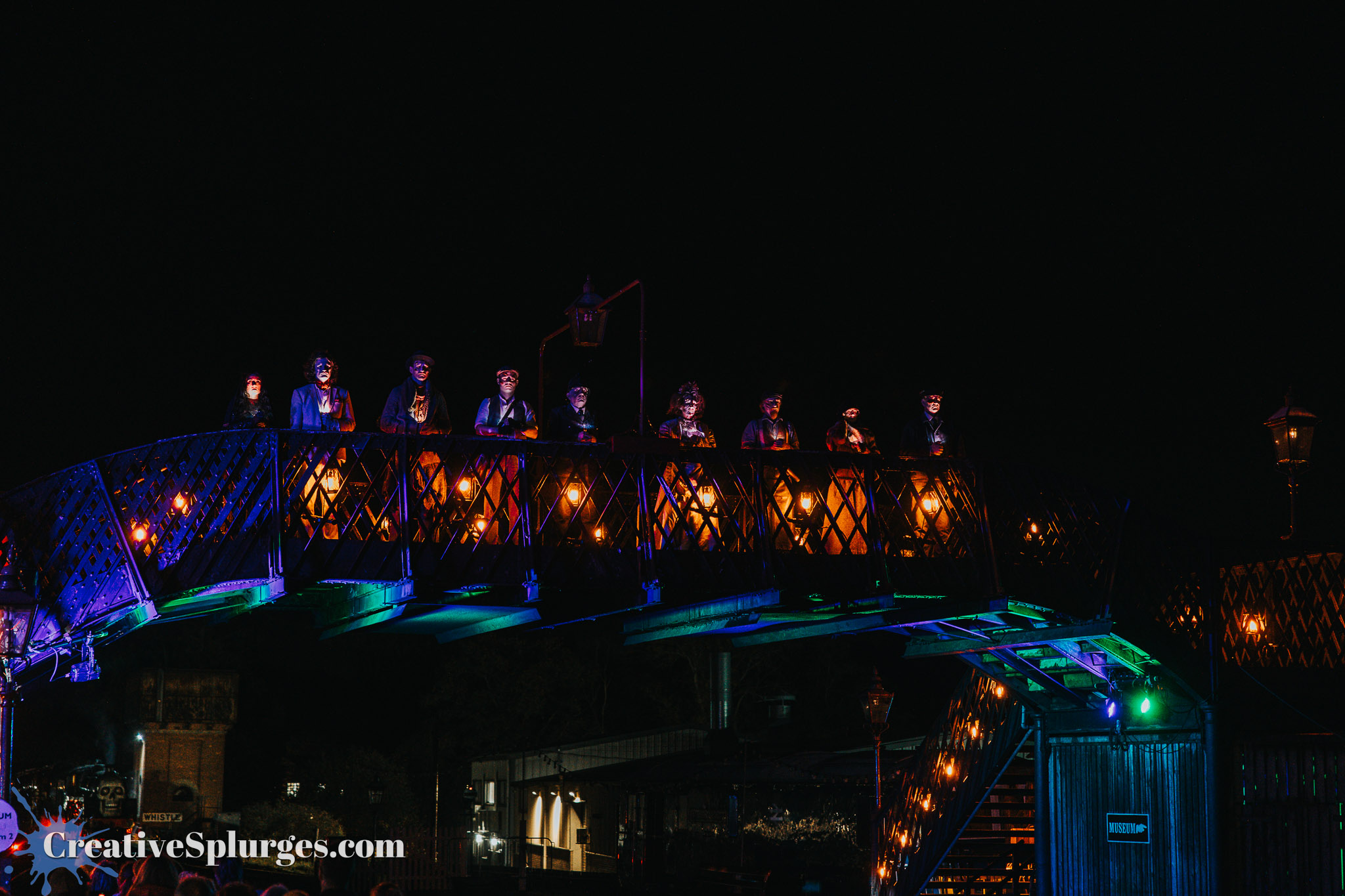A brightly lit bridge at night with figures dressed in various costumes standing along the railing, surrounded by glowing lanterns.