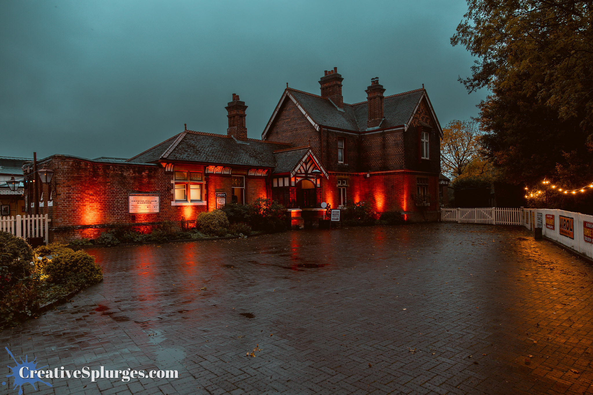 A historic brick building illuminated with orange lights on a rainy evening, surrounded by greenery and a white picket fence.