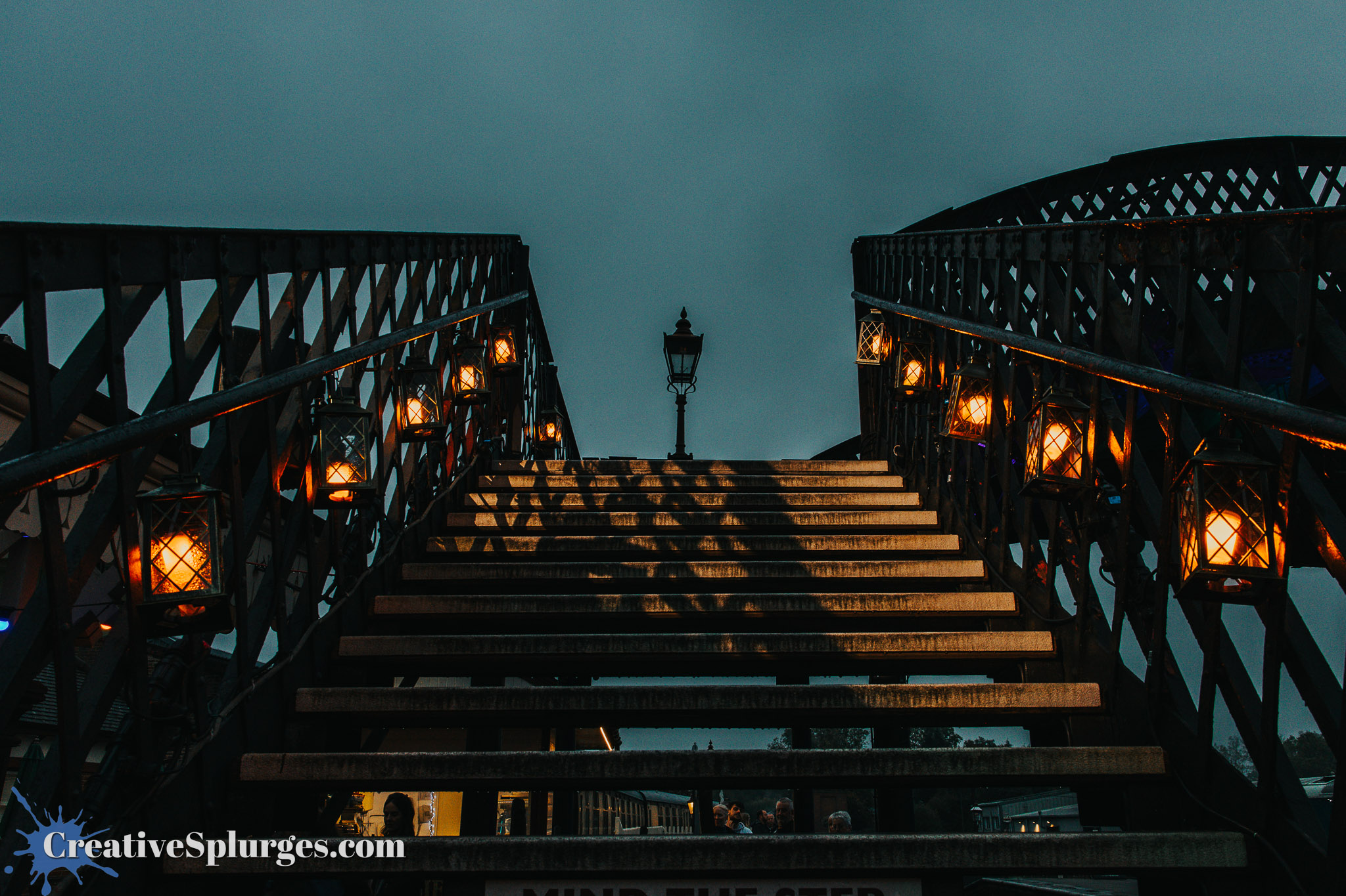 View of a staircase illuminated by lanterns, leading upward in an evening setting with a cloudy sky.