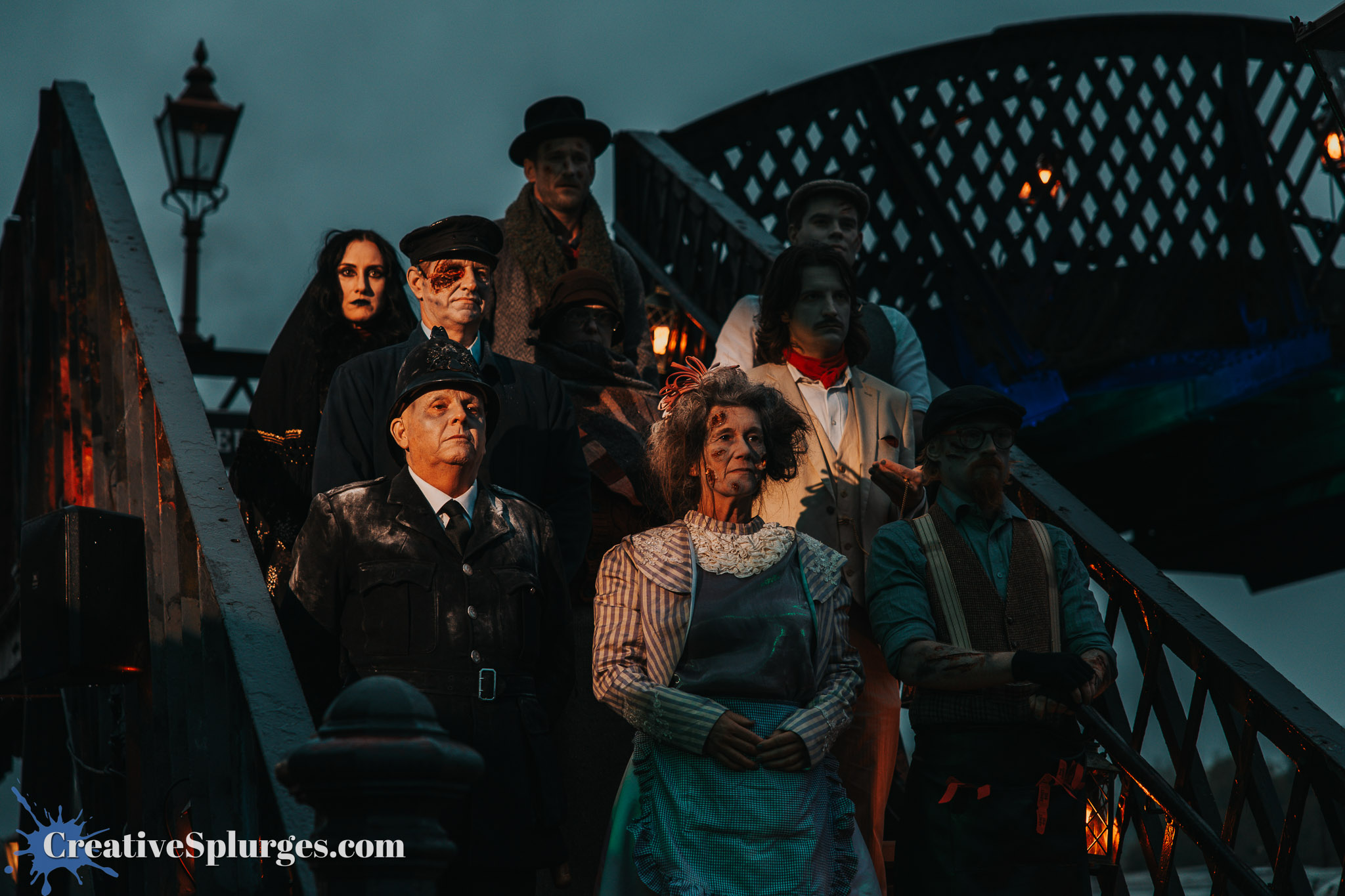 A group of individuals dressed in vintage, spooky costumes, standing on a staircase under a dim, overcast sky. The scene includes various characters with dark makeup and elaborate outfits, portraying a haunting or theatrical theme.