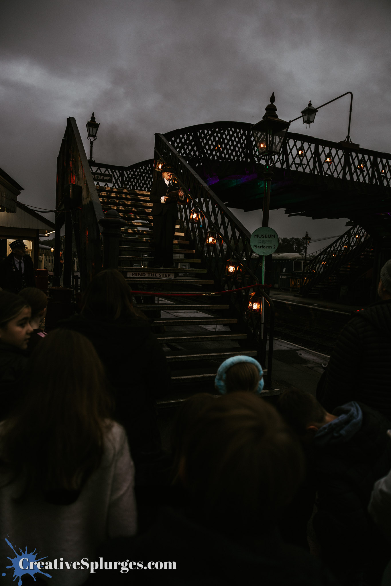 A crowd of children watches a performer in a suit standing on a staircase, illuminated by lamps, under a cloudy sky at a museum platform.