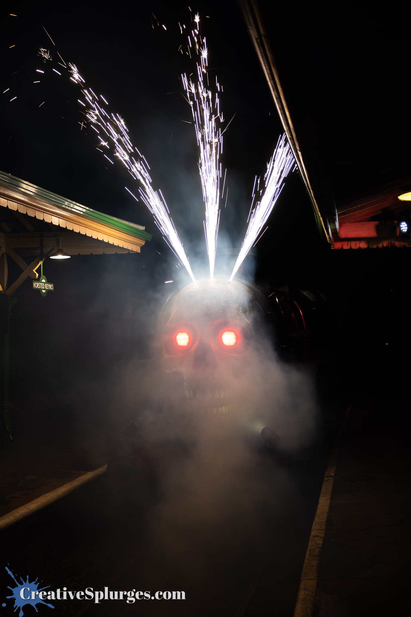 A large skull-shaped steam engine emits sparks and smoke, with glowing red eyes, at a train station at night.