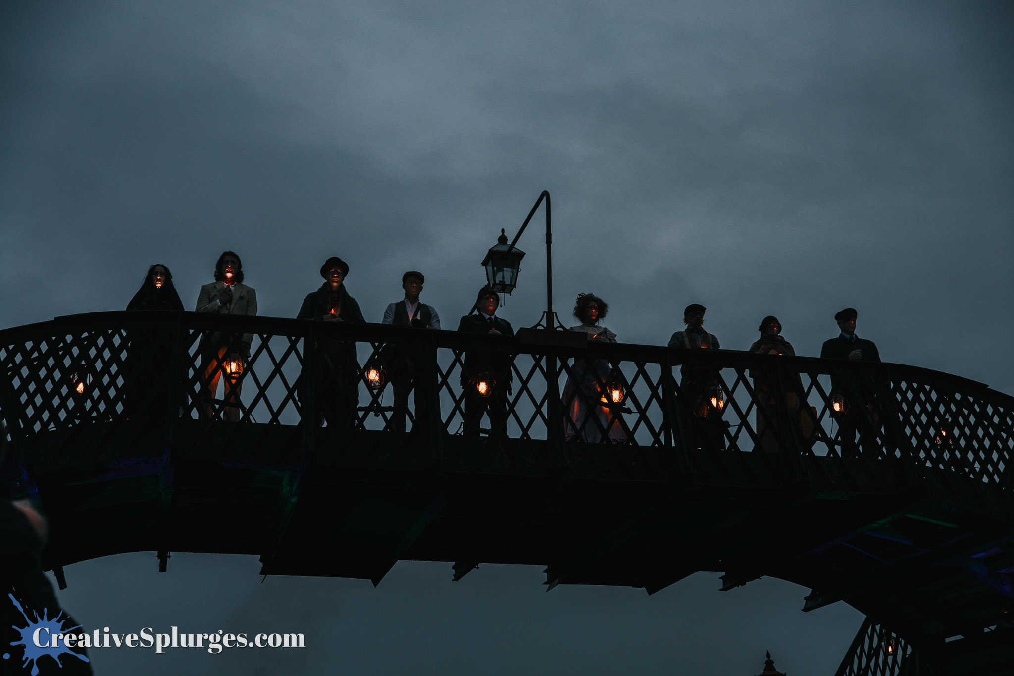 Group of people in period costumes holding lanterns, standing on a bridge against a dark, cloudy sky.