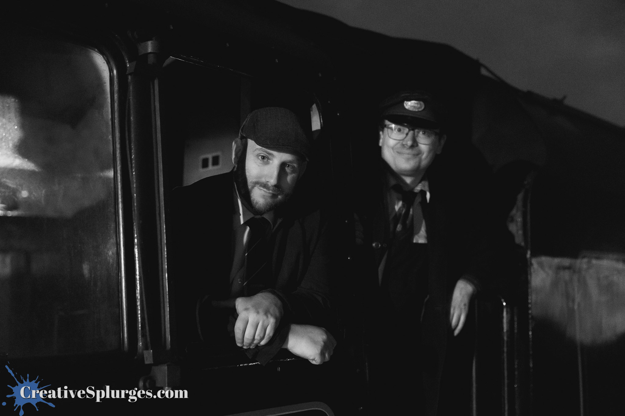 Two men in vintage attire pose inside a steam engine's cabin, one smiling and the other looking thoughtfully out the window, with a moody black and white backdrop.