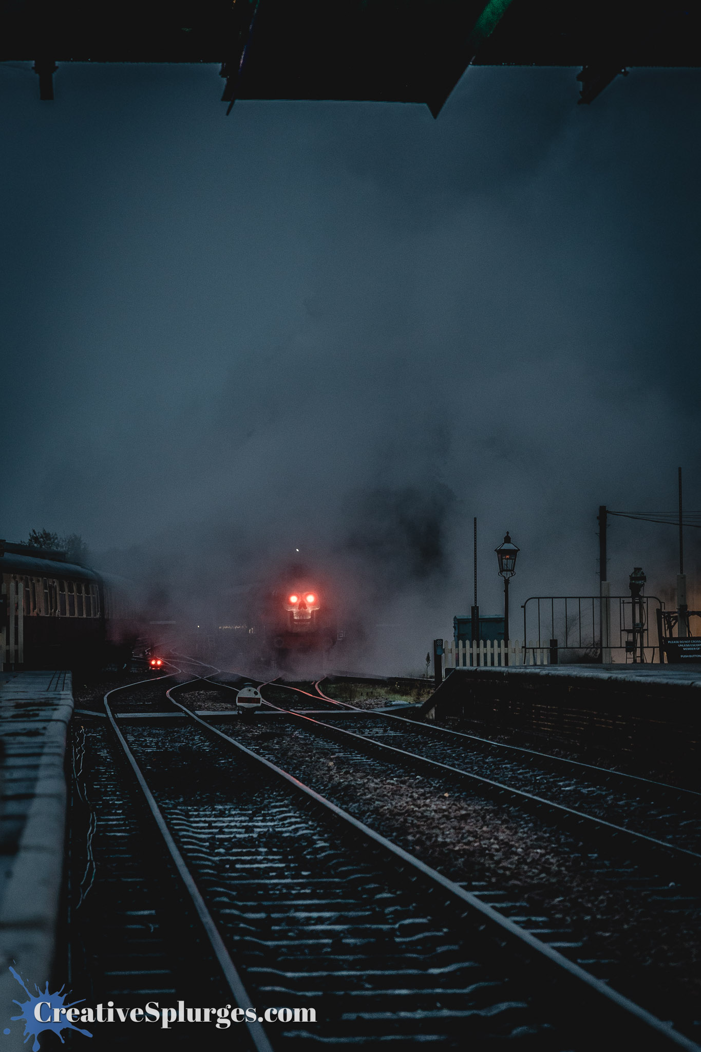 A steam train emerging from fog at a railway station, with glowing red lights and tracks curving away into the distance.