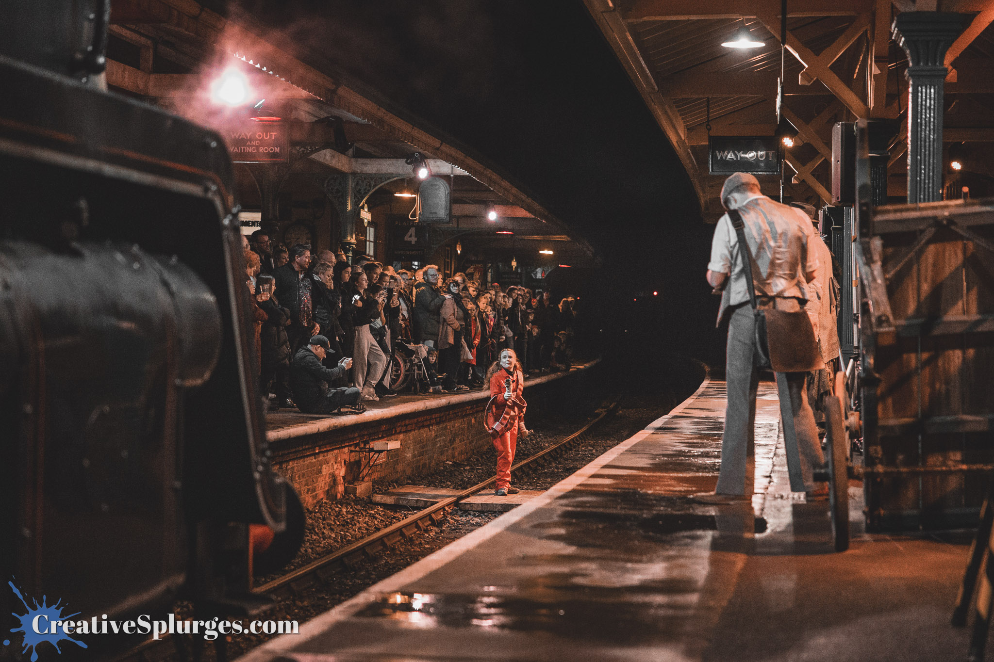 A crowd gathers at a dimly lit train station platform at night, watching a performance. A performer dressed in red stands on the tracks, while another performer in period attire stands near a wooden cart. Steam from an old steam locomotive billows in the foreground.