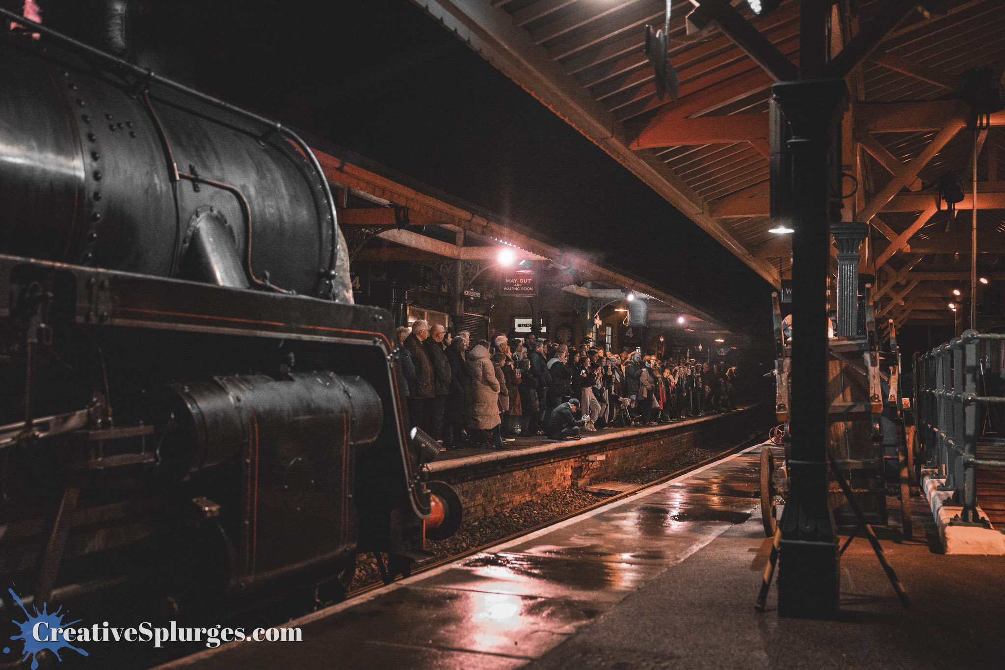 A vintage steam locomotive parked at a dimly lit train station, with a large crowd of people gathered on the platform, reflecting on the wet ground.