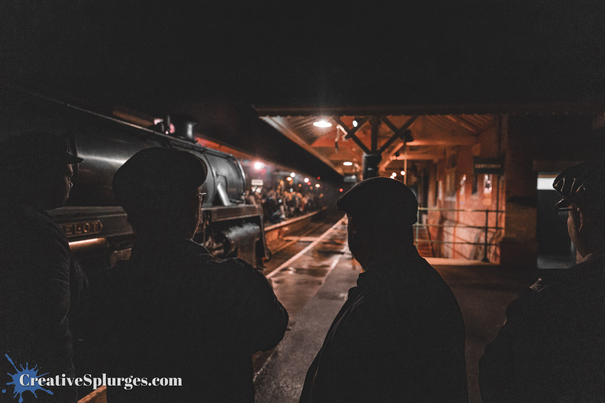 Silhouettes of two people in hats watching a steam locomotive at night, with a dimly lit train station in the background.