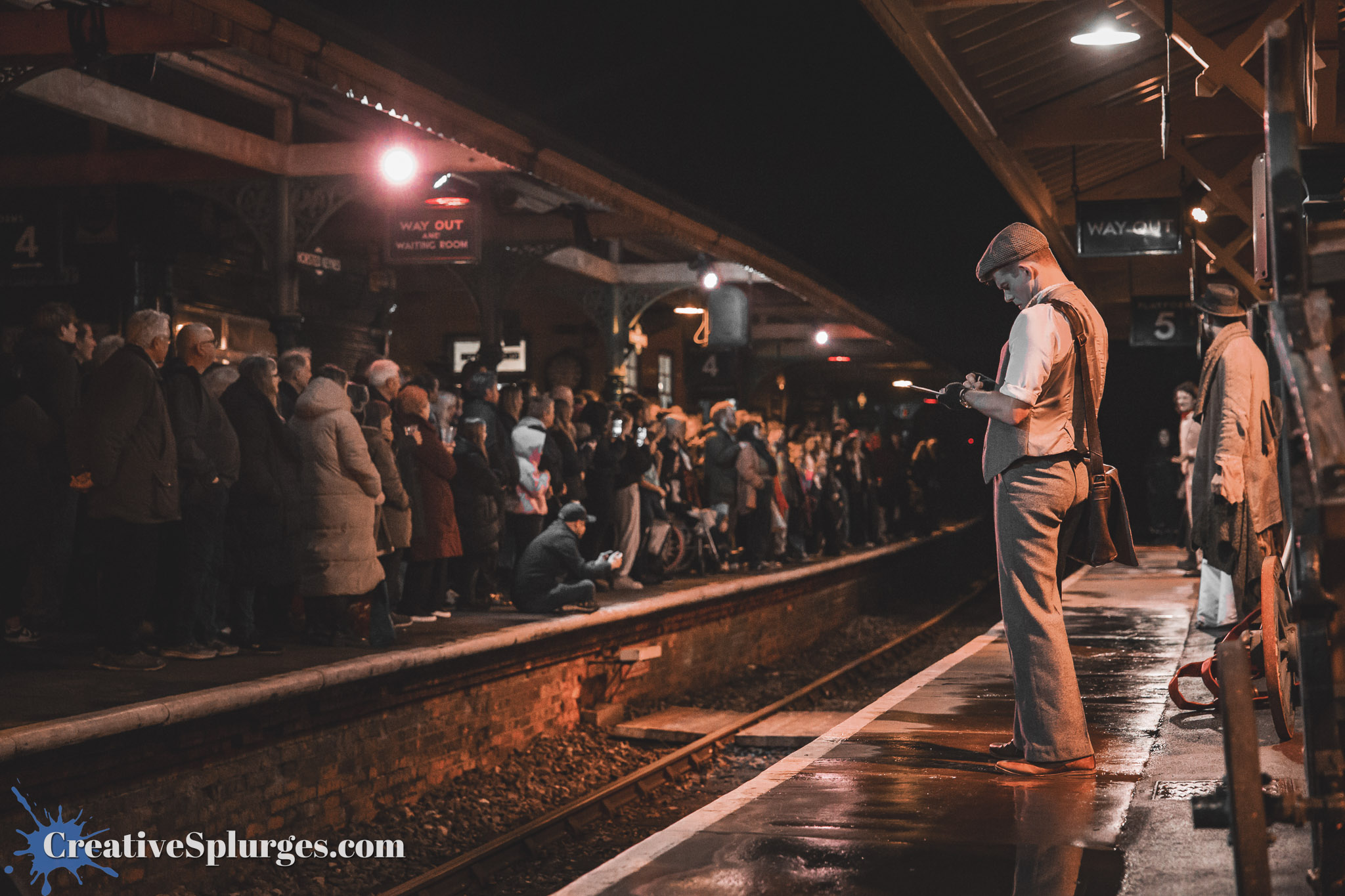 A crowded train platform at night, with a large group of people waiting, while a man in vintage attire stands in the foreground holding a device.