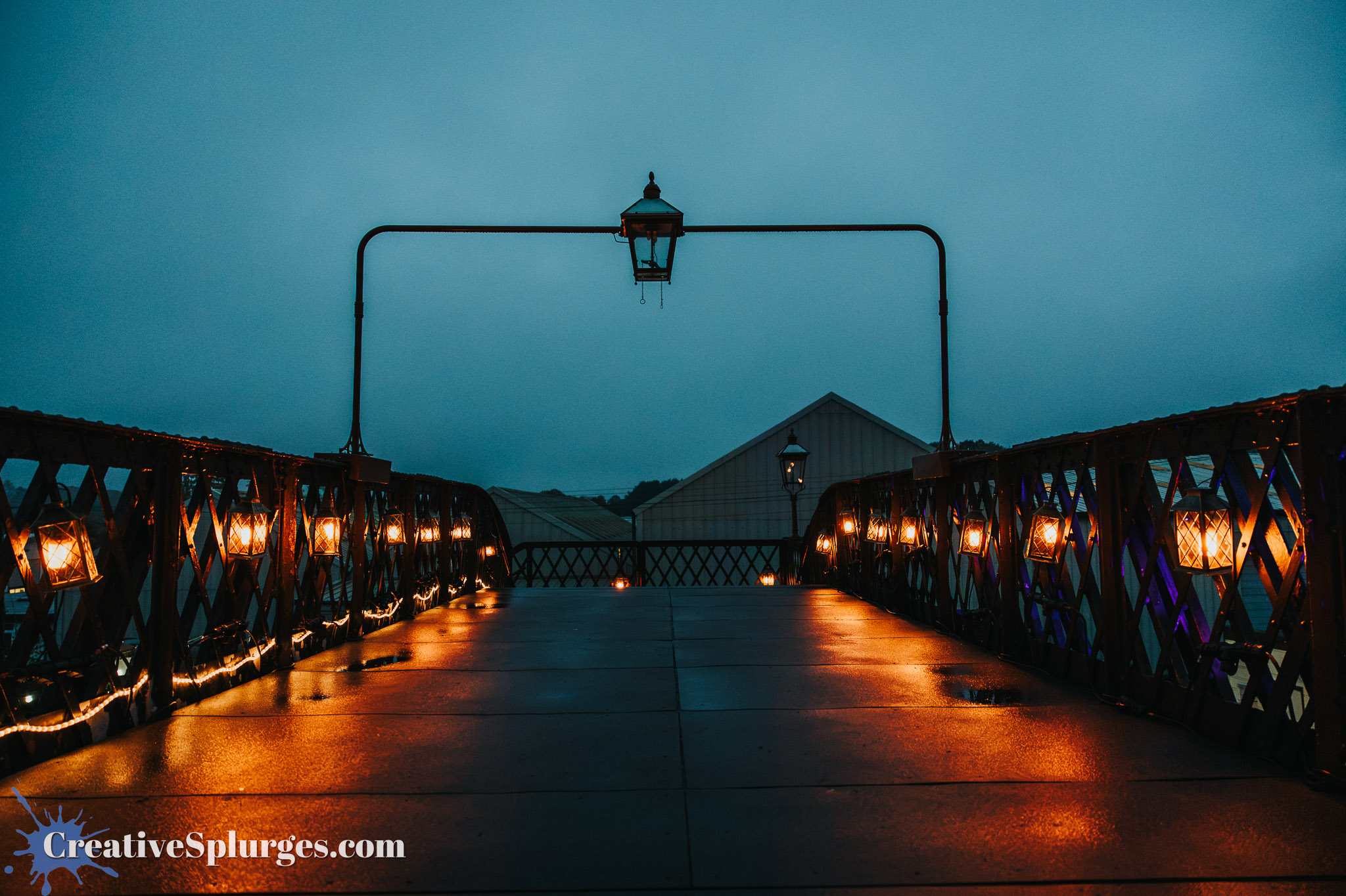 View of a bridge at dusk with decorative lanterns lighting the pathway and a cloudy sky in the background.