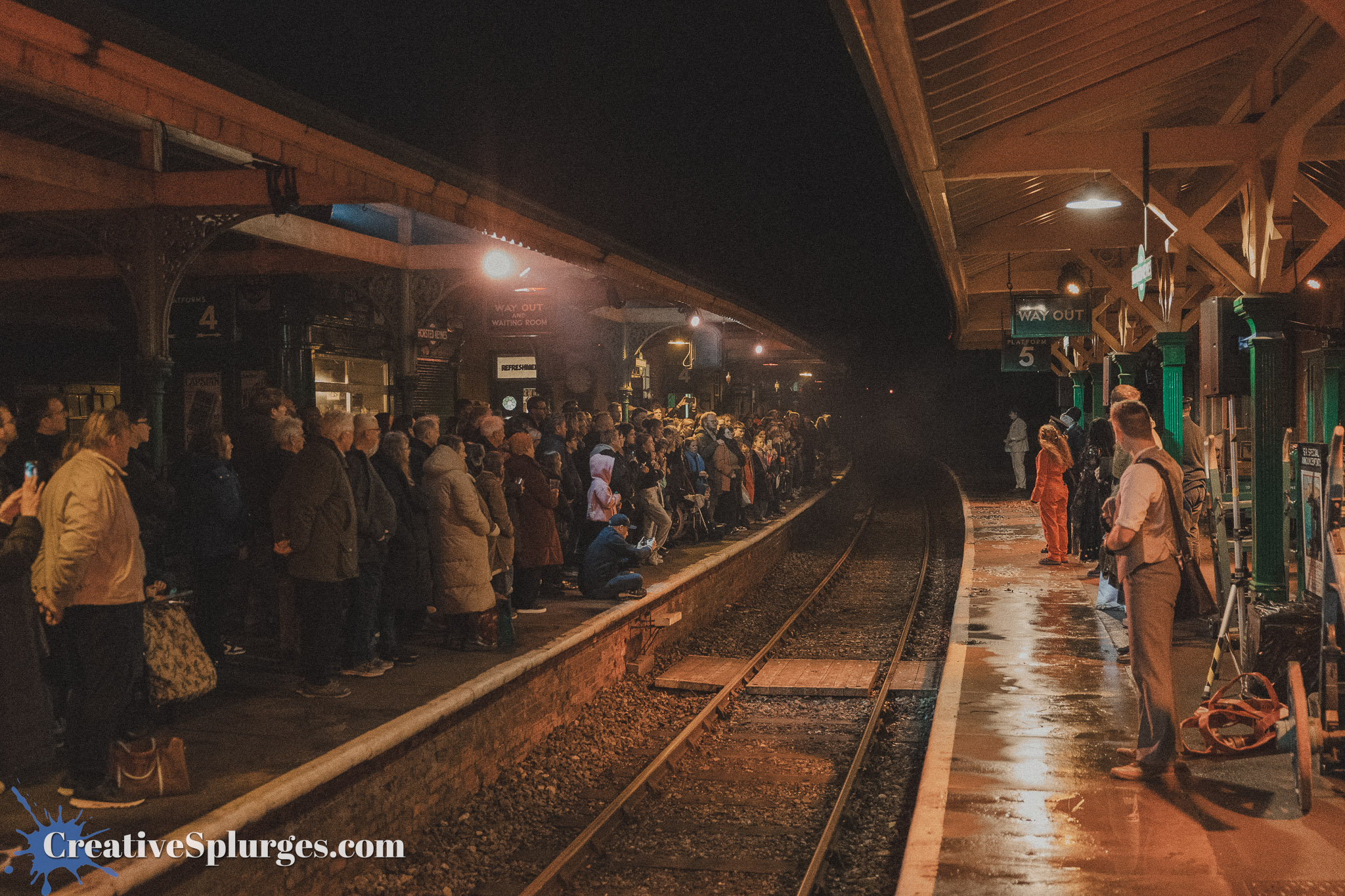 A crowded train station platform at night with a diverse group of people waiting. Some are standing, while others are seated. Train tracks run alongside the platform, and multiple signs and lighting indicate the station amenities.