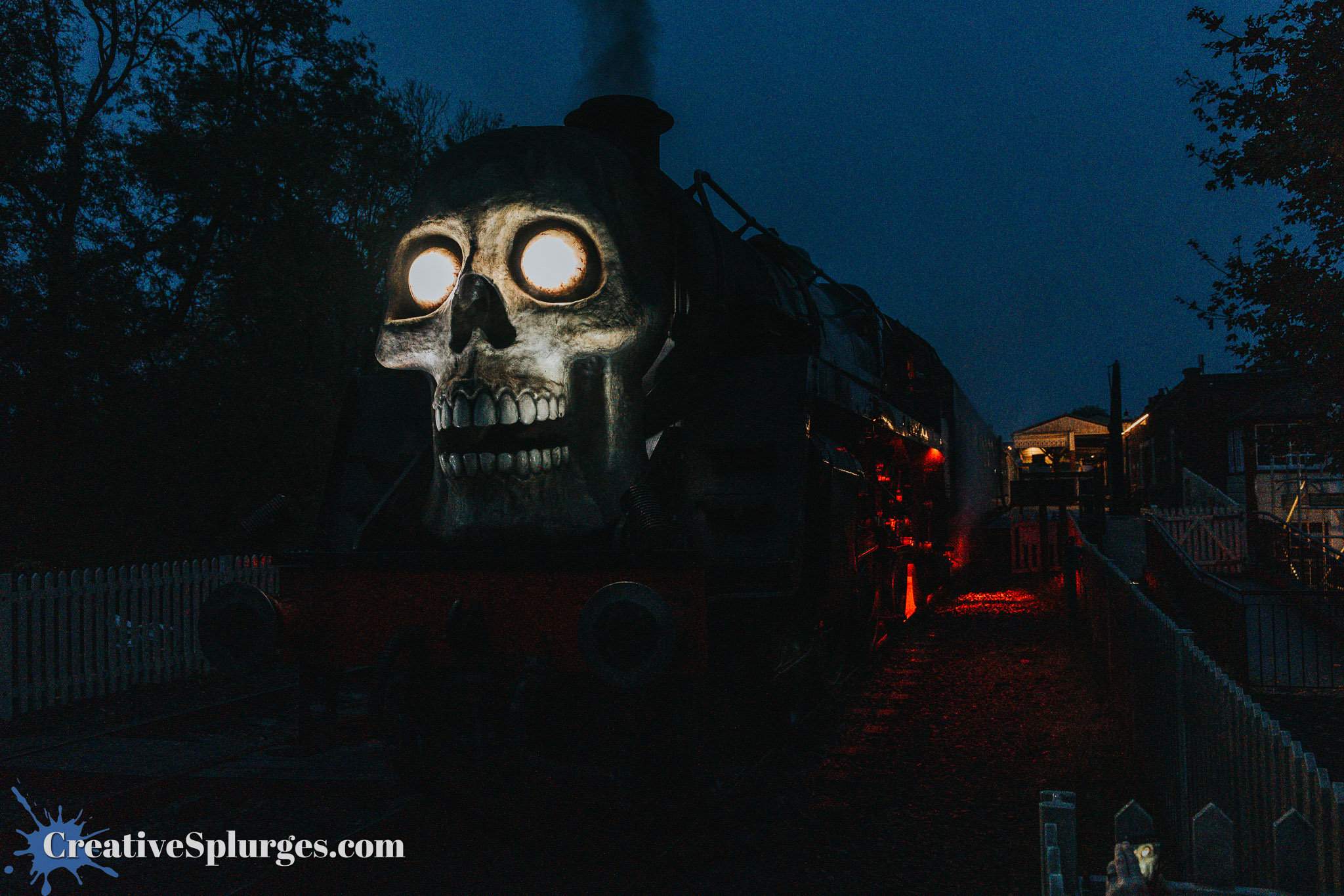 A steam locomotive with a skull design on the front, illuminated in the dark with glowing eyes, surrounded by trees and a white picket fence.