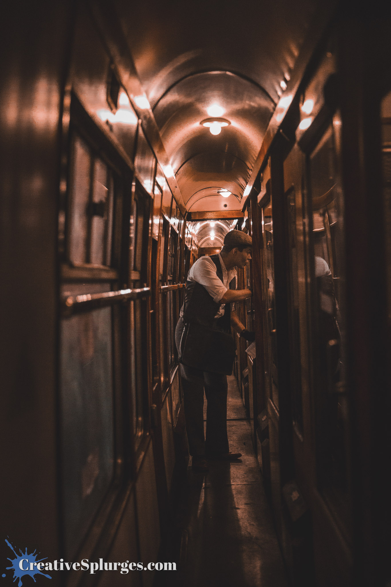 A man in vintage attire standing in a narrow train corridor, inspecting a window. The interior is dimly lit with a warm glow from overhead lights, with wooden panels and glass windows lining the walls.