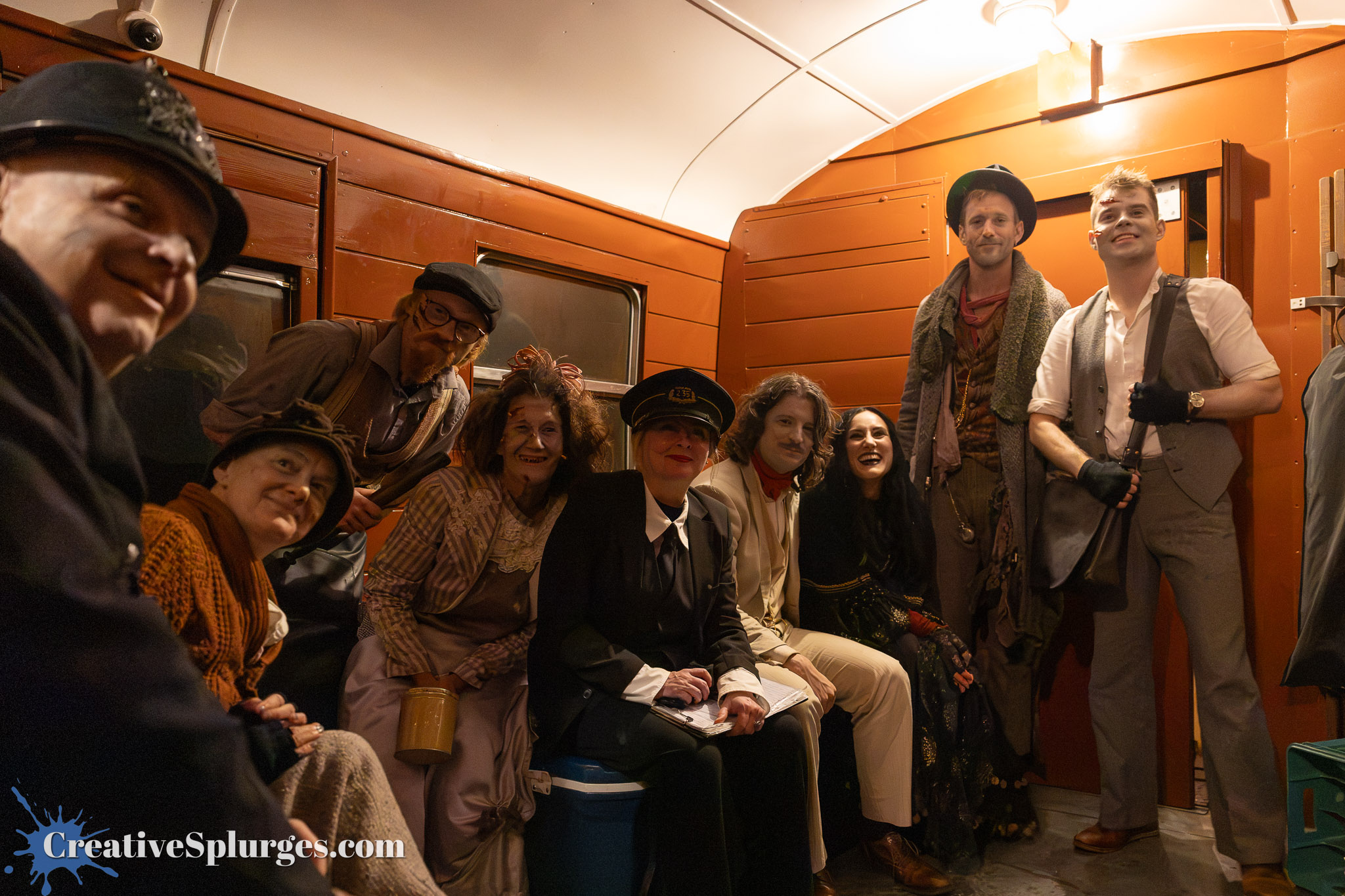 A group of actors in costumes, seated inside a vintage train carriage, smiling and posing for the camera.