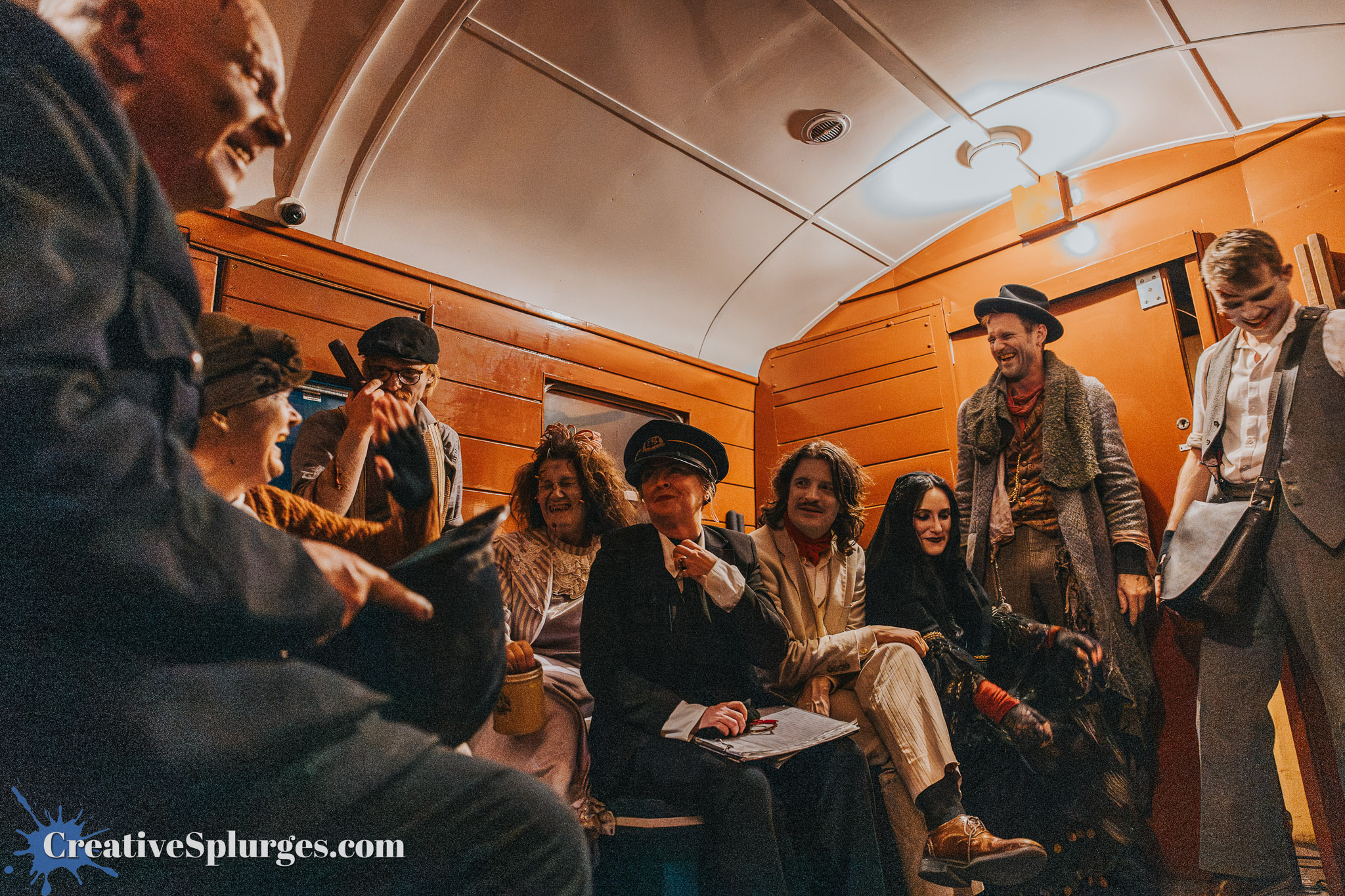 A group of people dressed in vintage costumes sitting together in a colorful interior, likely of a train or vintage carriage, with some laughing and interacting.