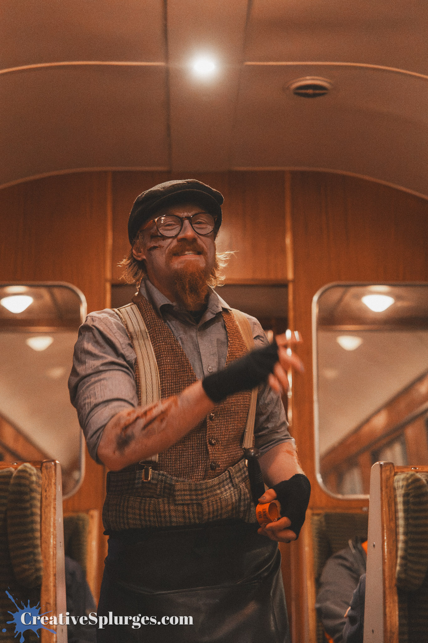 A bearded man in vintage attire, including a cap and suspenders, stands inside a train carriage, smiling while holding a small object, with some makeup resembling scars on his face.