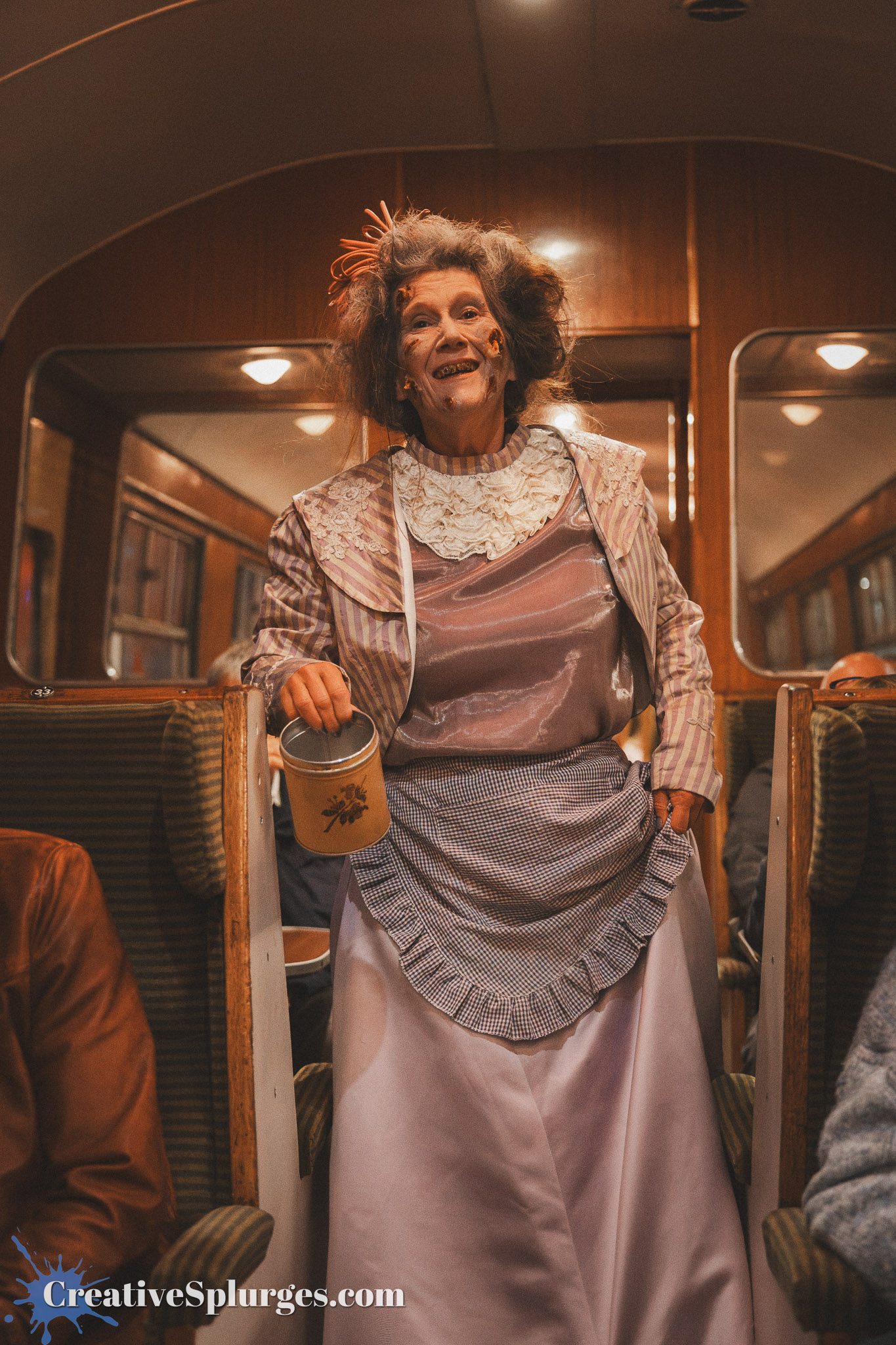 A person dressed in a vintage-style costume with exaggerated makeup depicting an elderly character, standing inside a train carriage, holding a container.