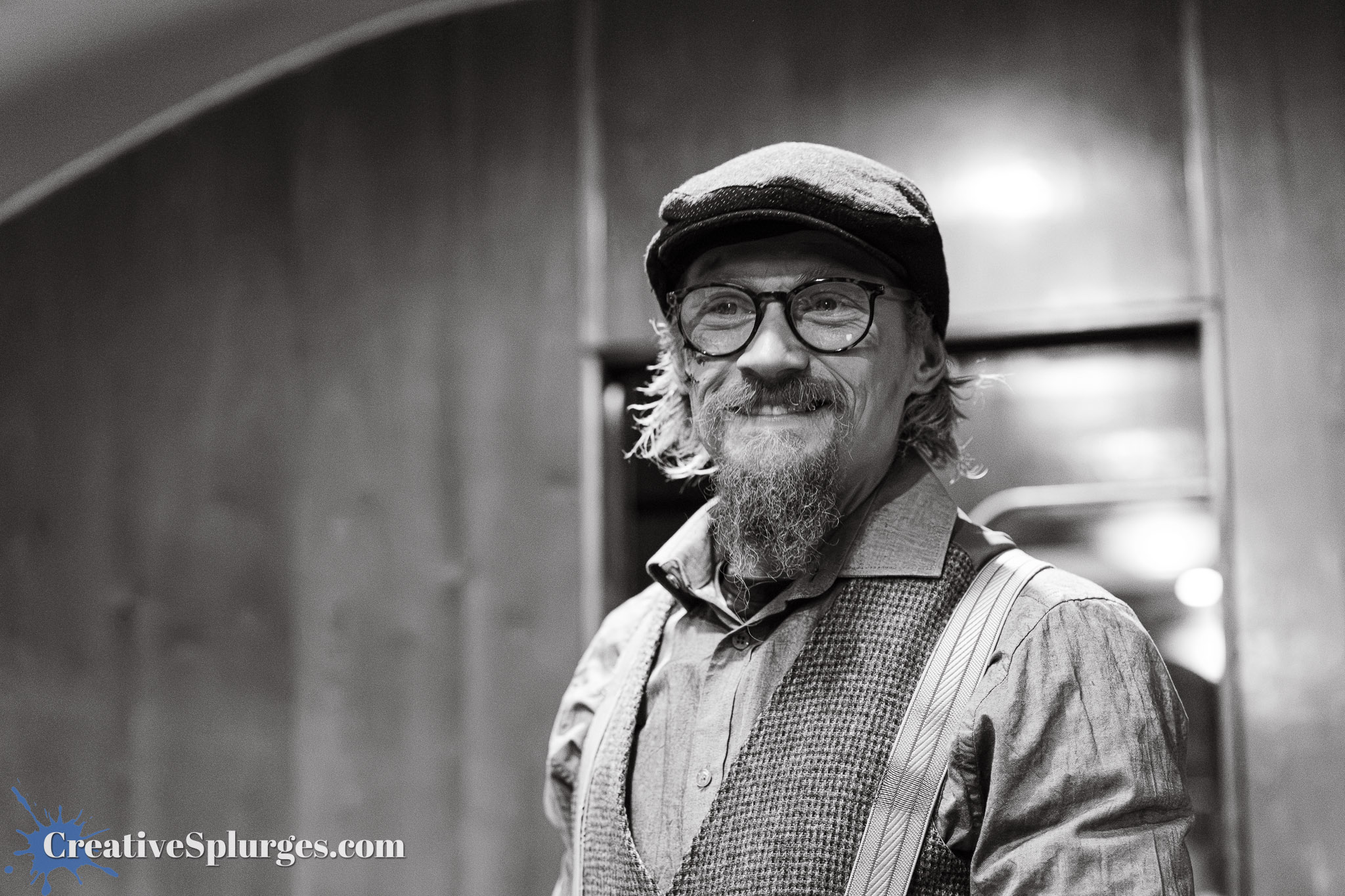 A smiling man with glasses and a flat cap, wearing a vest and suspenders, standing in a dimly lit interior.