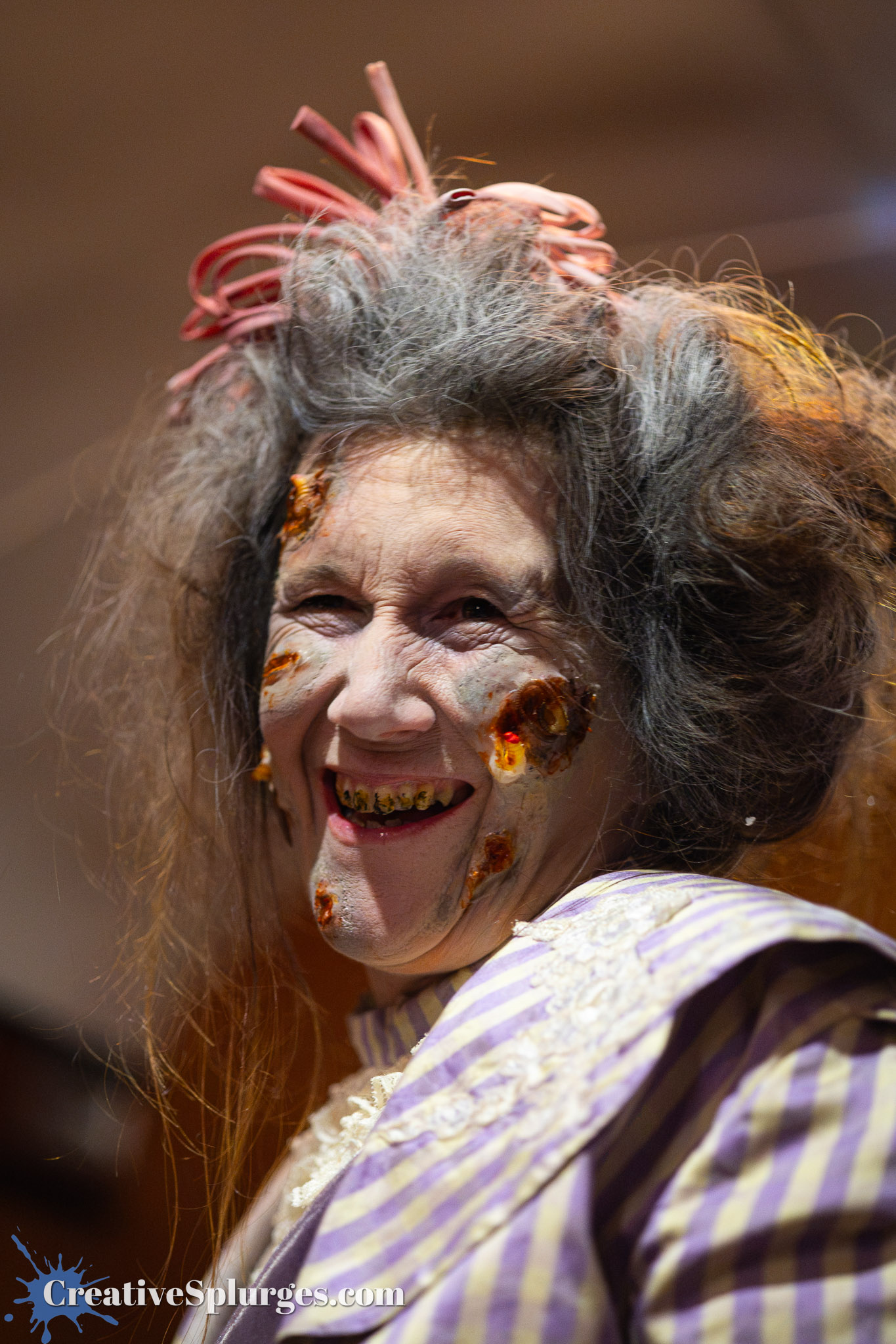 A person with dramatic makeup resembling a zombie, featuring discolored skin and rotting teeth, wearing a vintage striped outfit with lace detailing.
