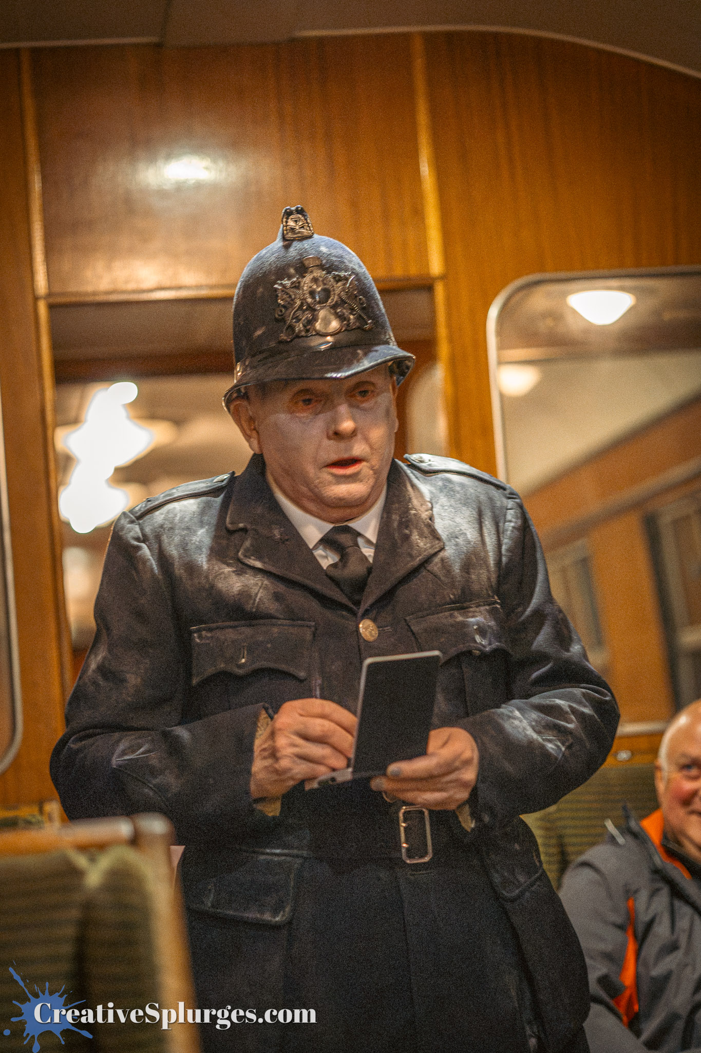 A performer dressed as a vintage police officer in a black uniform and helmet, holding a notebook, inside a train carriage with wooden paneling.