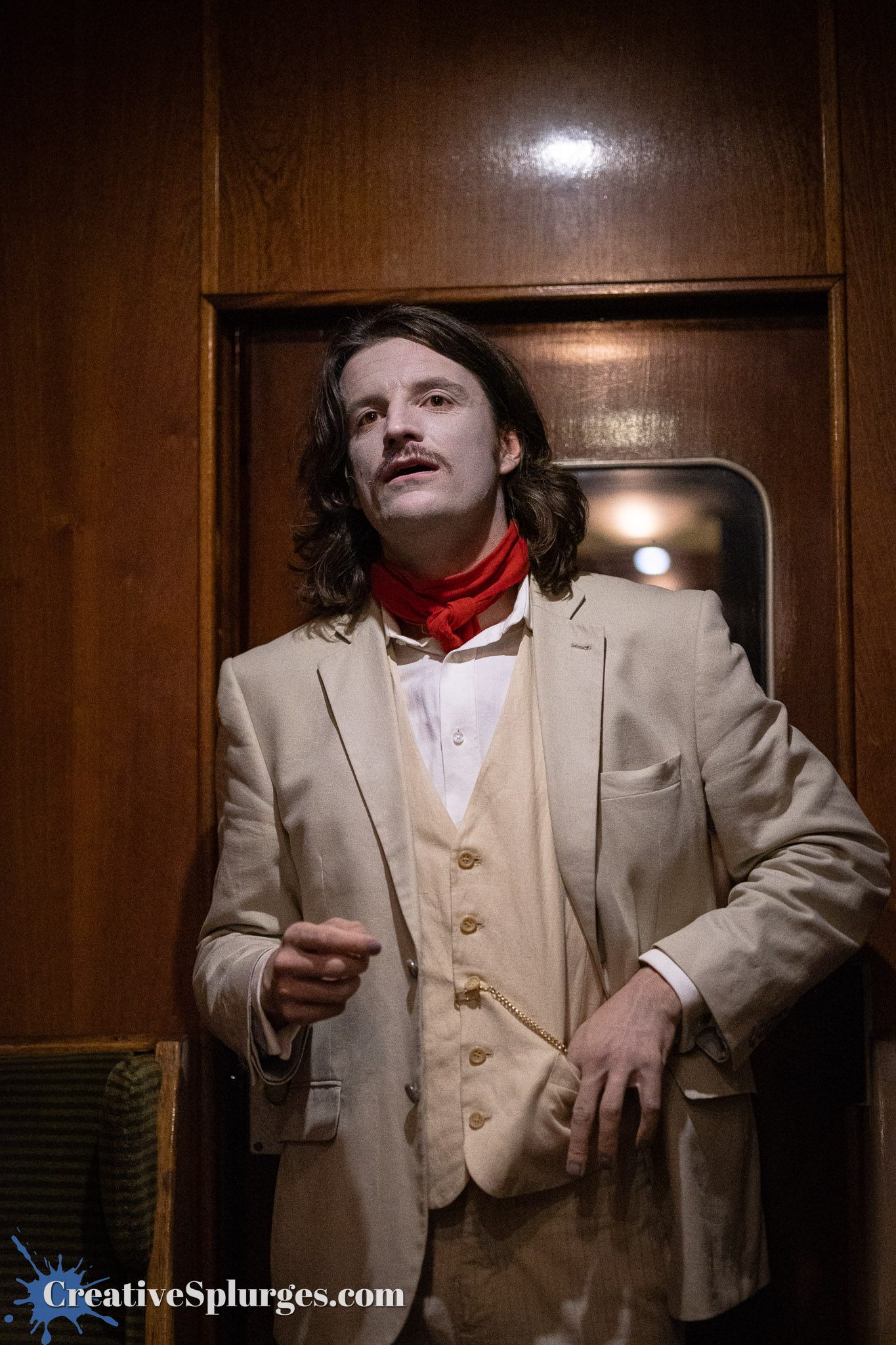A man dressed in a vintage beige suit with a red scarf, standing inside a wooden interior, holding a pose and looking slightly to the side.