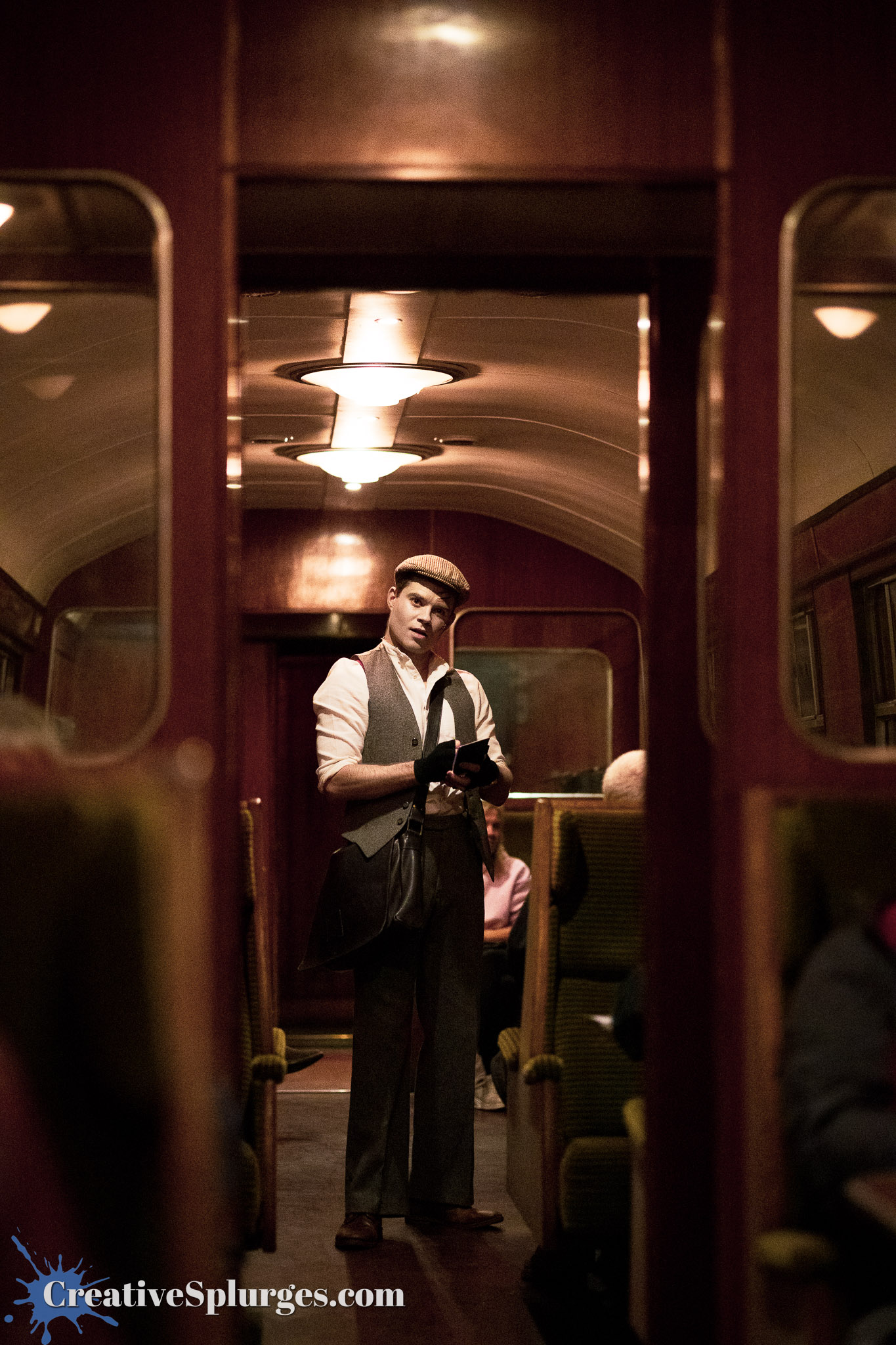 A man in period clothing stands in an old-fashioned train carriage, holding a book and looking at the camera. The interior features wooden paneling and retro lighting, with passengers seated in the background.