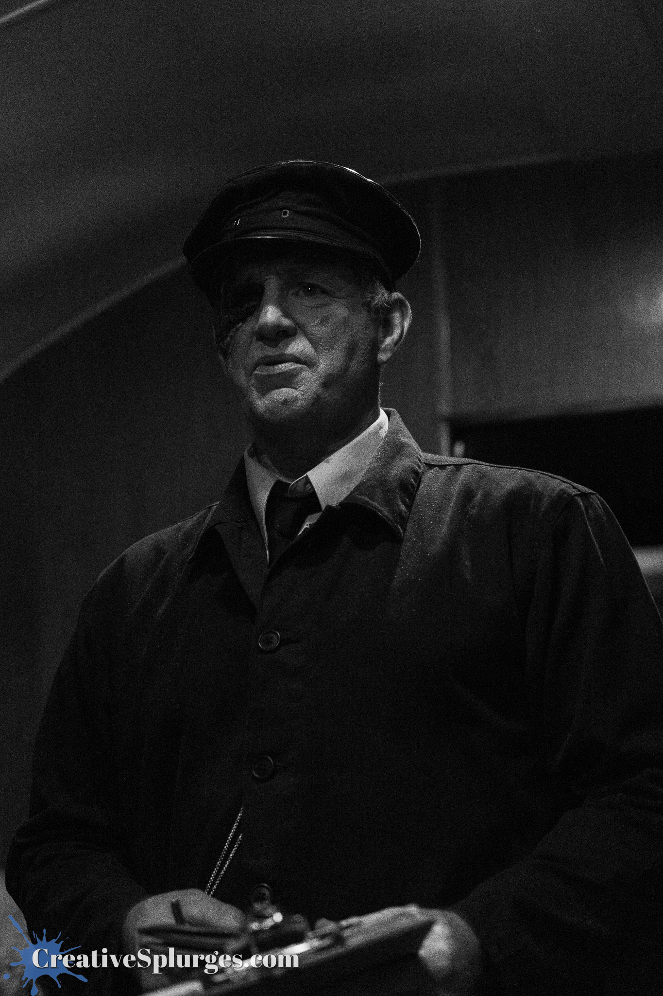 A man in a black coat and cap, with a visible facial scar, stands holding a clipboard in a dimly lit setting.