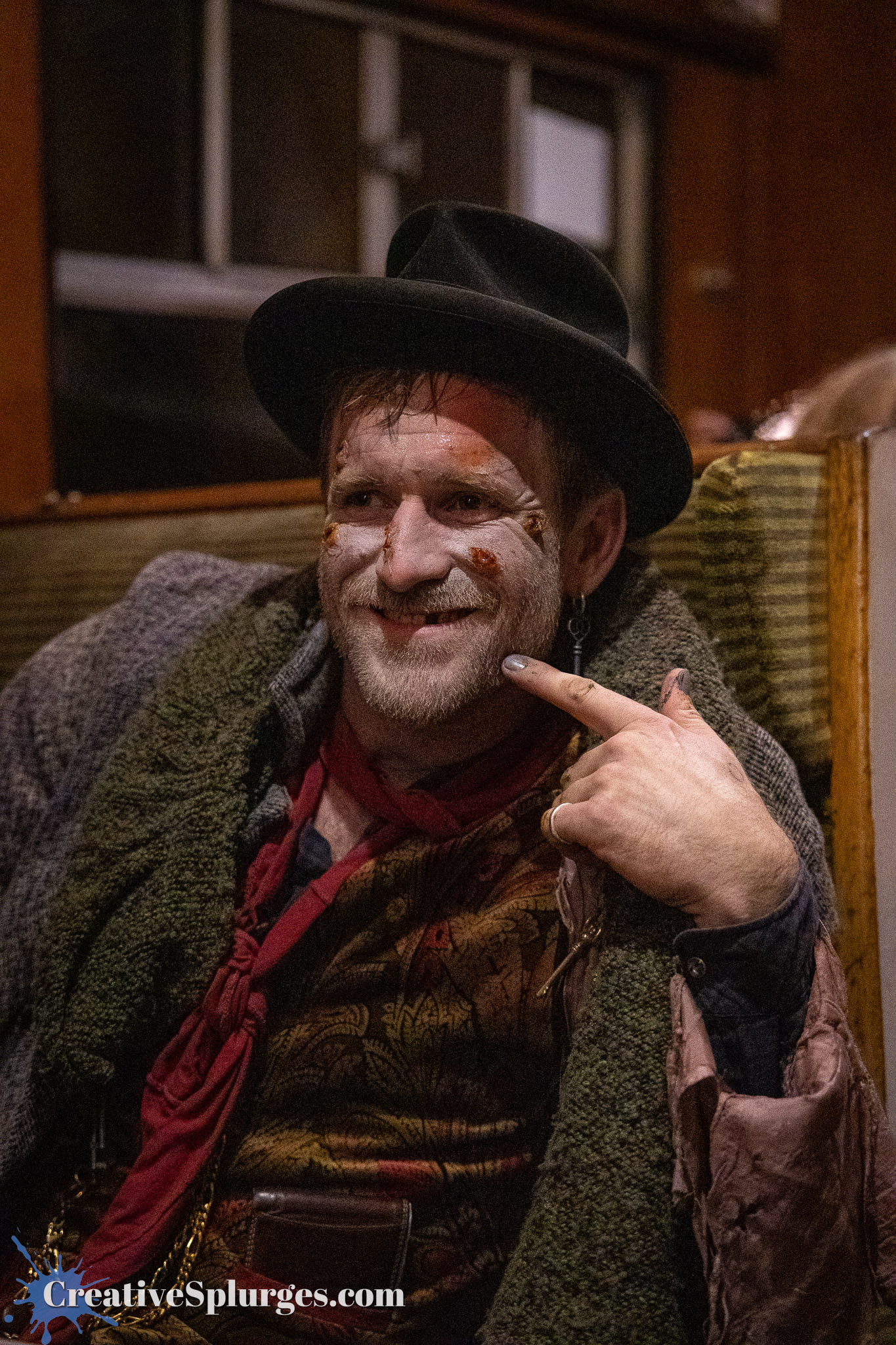 A man with a playful expression, wearing a black hat and colorful layered clothing, with distinct makeup and facial embellishments, sitting indoors.