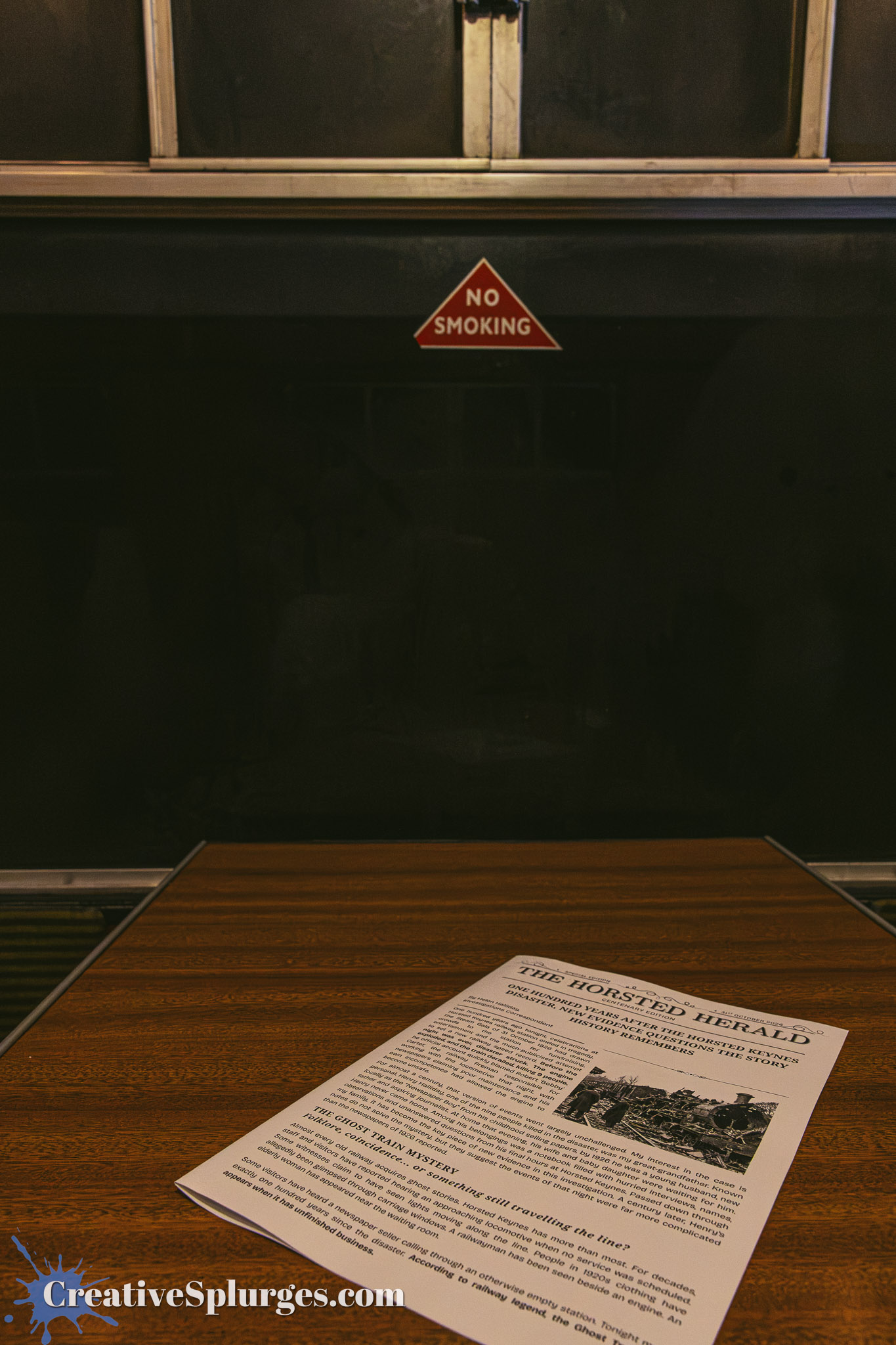 A wooden table with a newspaper titled 'The Horsted Herald' lying on it, featuring a large text and images related to history. In the background, a 'No Smoking' sign hangs on the wall.