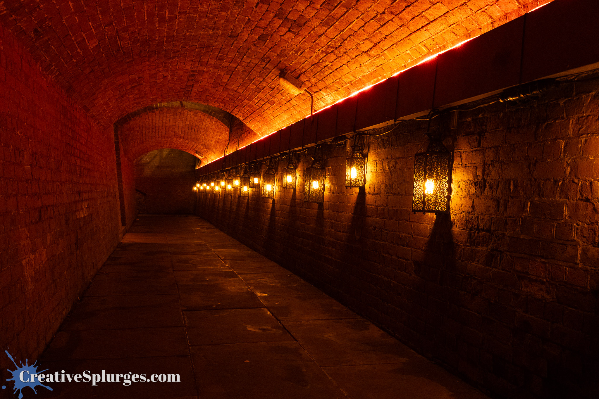 Dimly lit brick tunnel with arched ceiling and decorative lanterns lining the walls.