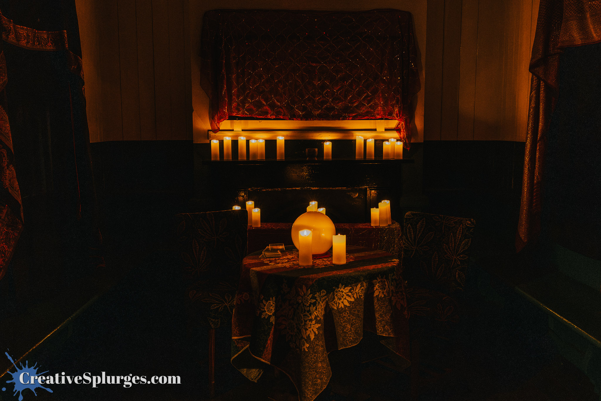 A dimly lit room featuring a small table covered with an ornate tablecloth, surrounded by chairs. The table holds a crystal ball and several lit candles, while more candles are arranged on a shelf in the background. Warm lighting creates a mystical atmosphere.