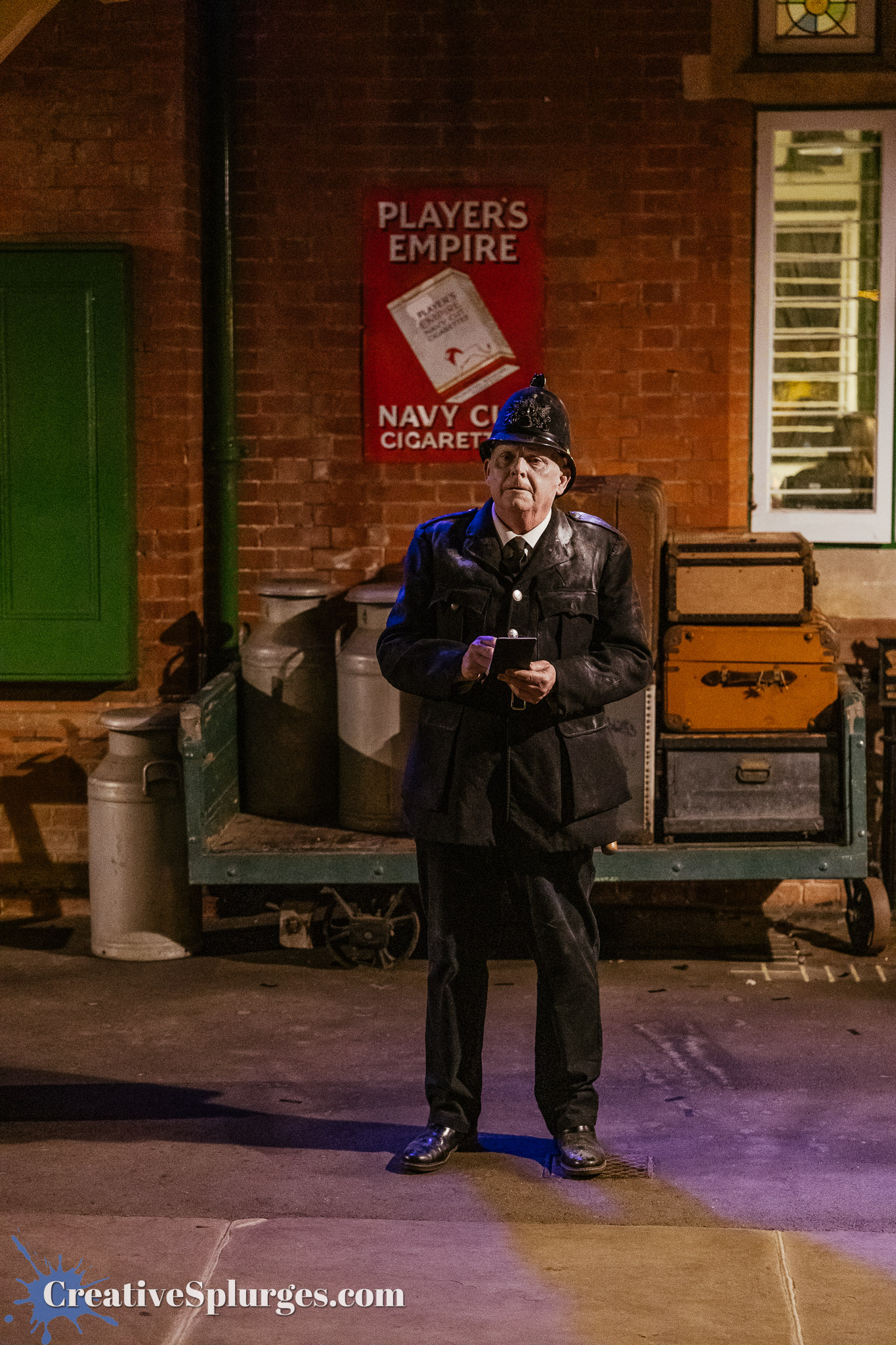 A vintage-style police officer standing on a stage, holding a notepad, with a brick wall and old luggage in the background. A sign for 'Player's Empire' and 'Navy Cut Cigarettes' is visible on the wall.