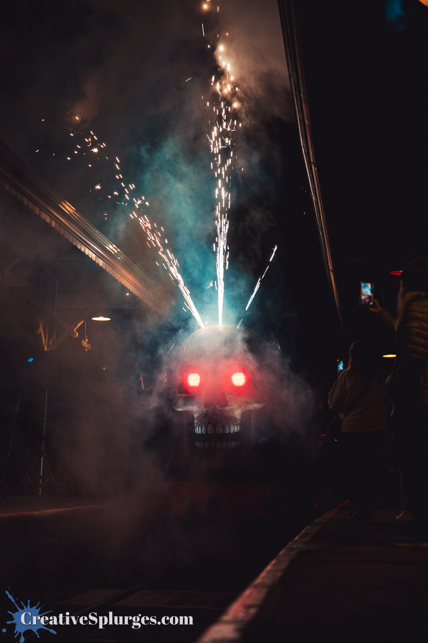 A spooky train with a skull face and glowing red eyes emerges from smoke, surrounded by sparks and fog, while people watch and take photos from the platform.