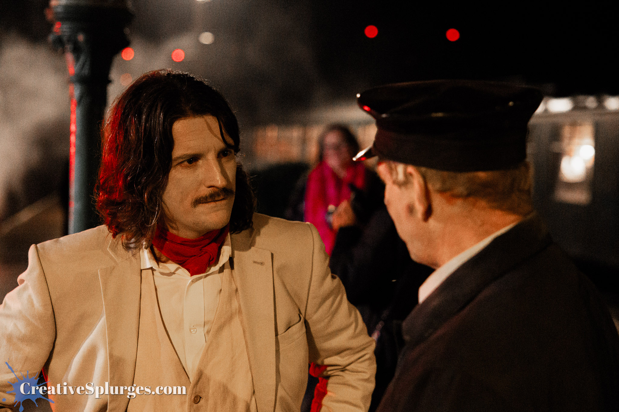 Two men engaged in a serious conversation at a nighttime outdoor location, one dressed in a light suit with a red scarf and the other in a dark coat and cap, with a train in the background.