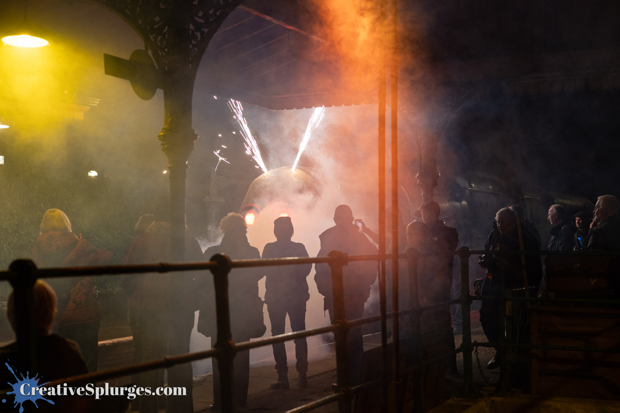 A group of people silhouetted against a smoky background watch a train with sparks emitting from it, creating a dramatic scene at a train station.