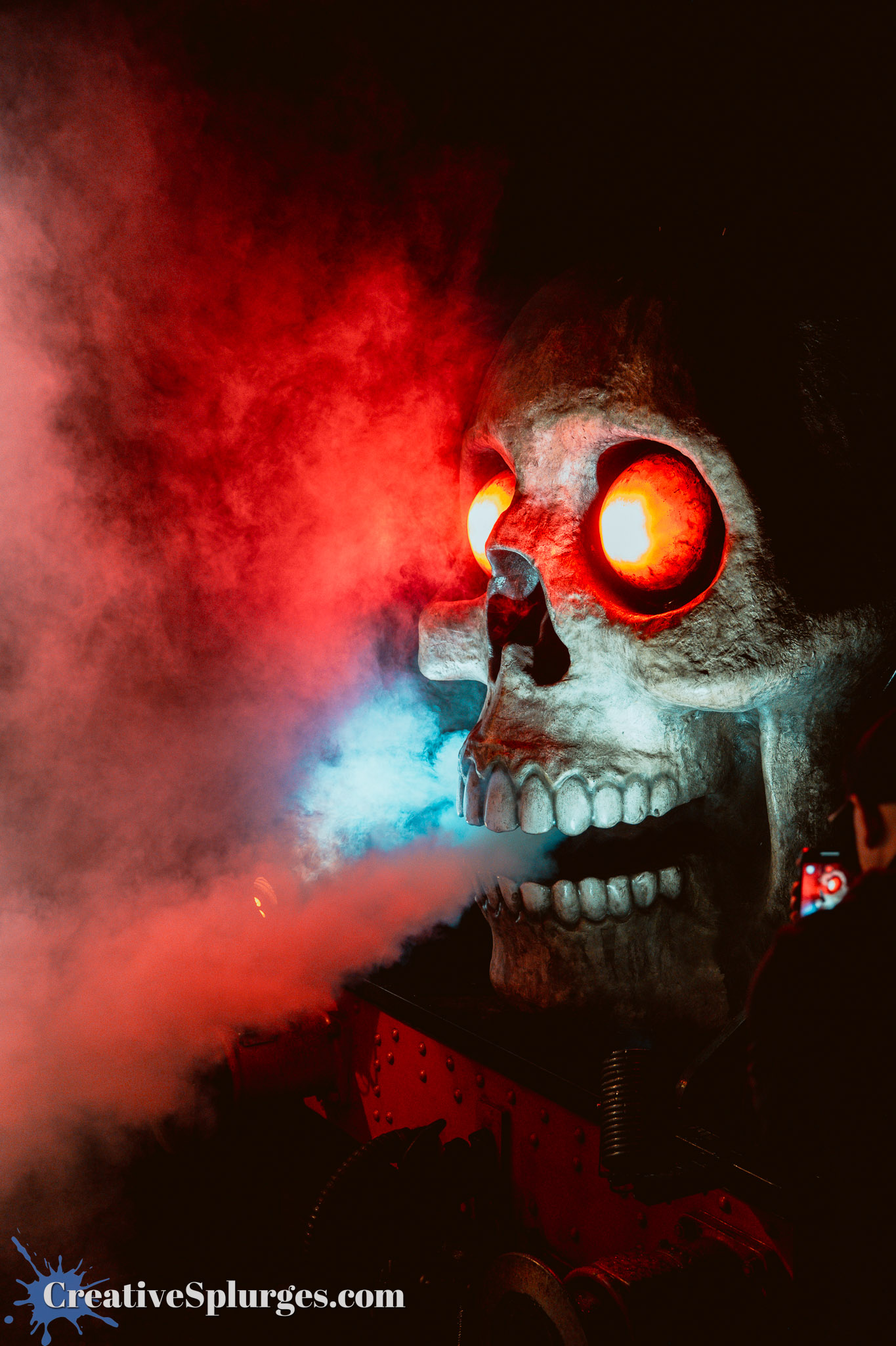 A large, illuminated skull with glowing red eyes emitting colored smoke, set against a dark background.