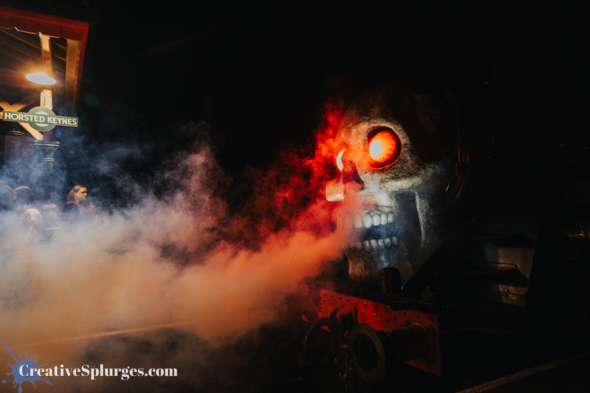A steam train with a skull-themed front, emitting smoke and illuminated red eyes, set against a night backdrop at Horsted Keynes station.