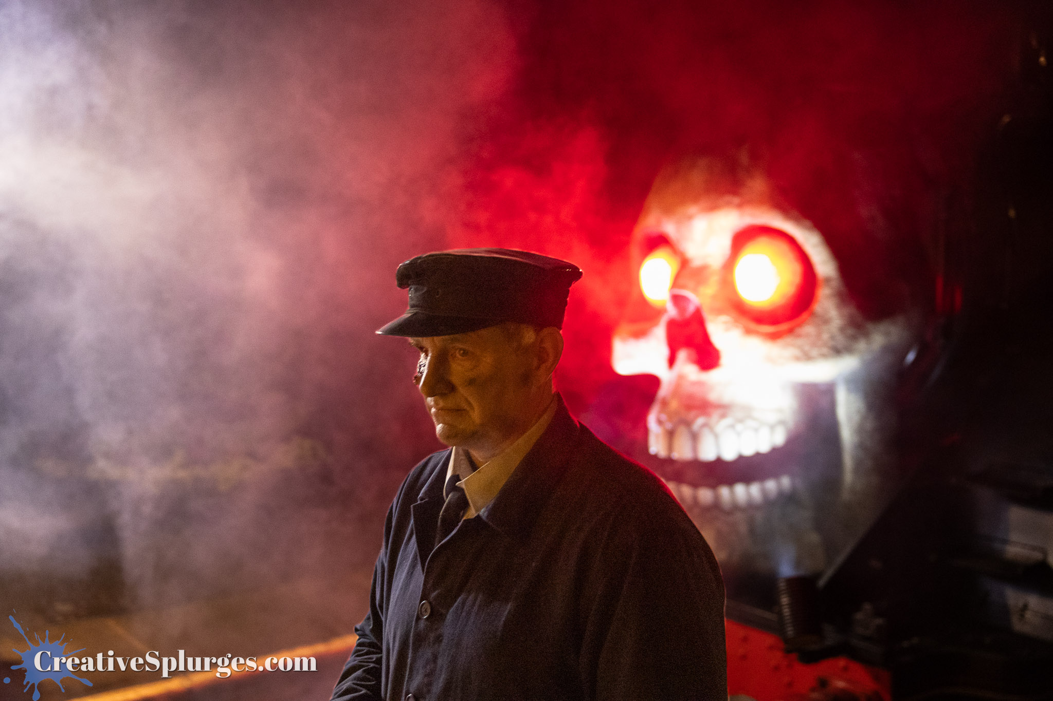 A person dressed in a dark coat and hat stands in front of a steam train, with a large skull illuminated behind them. The scene is enhanced by dramatic fog and red lighting, creating a spooky atmosphere.