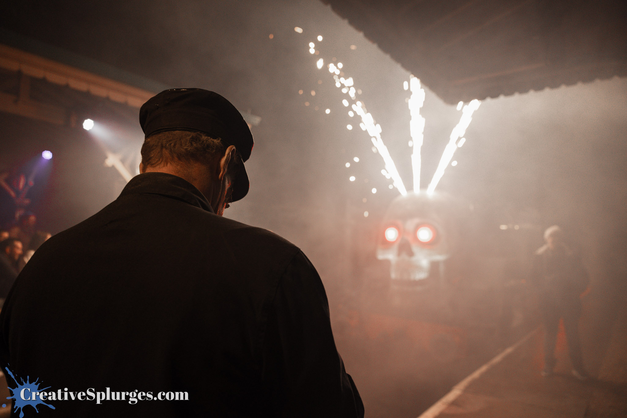 A man in a black cap and coat stands with his back to the camera, observing a scene with a train that has a large skull design and red glowing eyes, surrounded by smoke and fireworks.