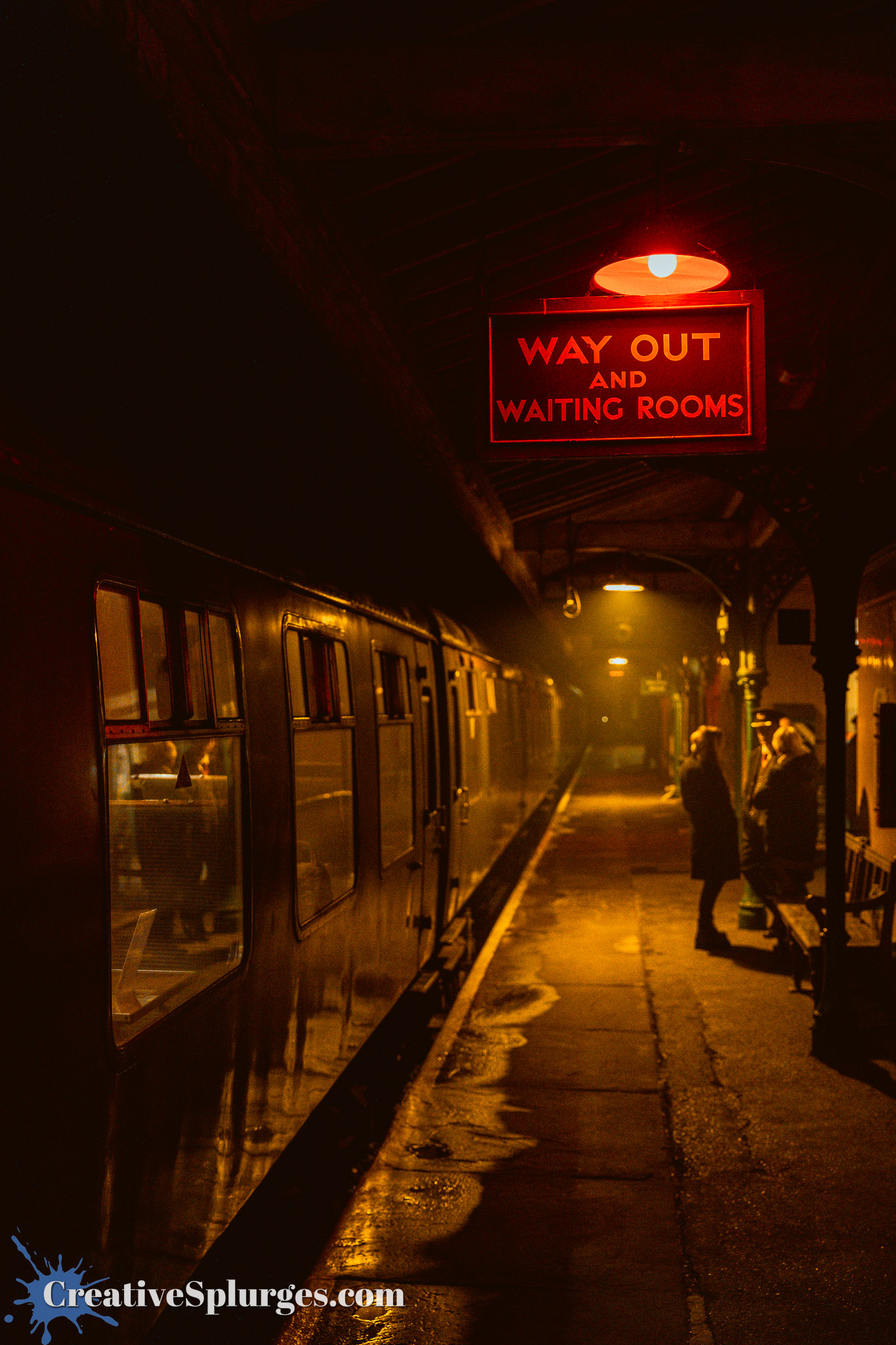 An illuminated sign reading 'WAY OUT AND WAITING ROOMS' hangs above a dark train platform with a vintage train in the background, creating a moody atmosphere.