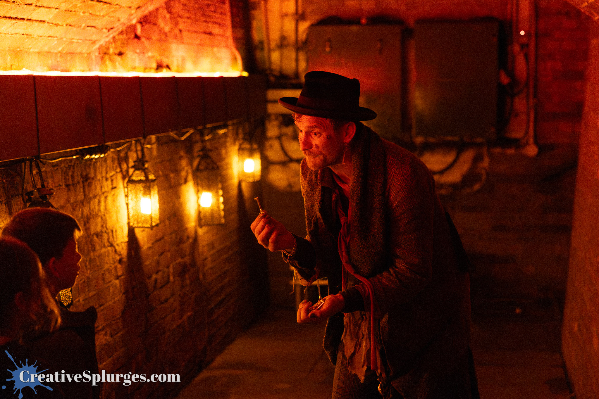 A man in a hat and coat, engaging with children in a dimly lit, atmospheric space with warm-toned lights and brick walls.